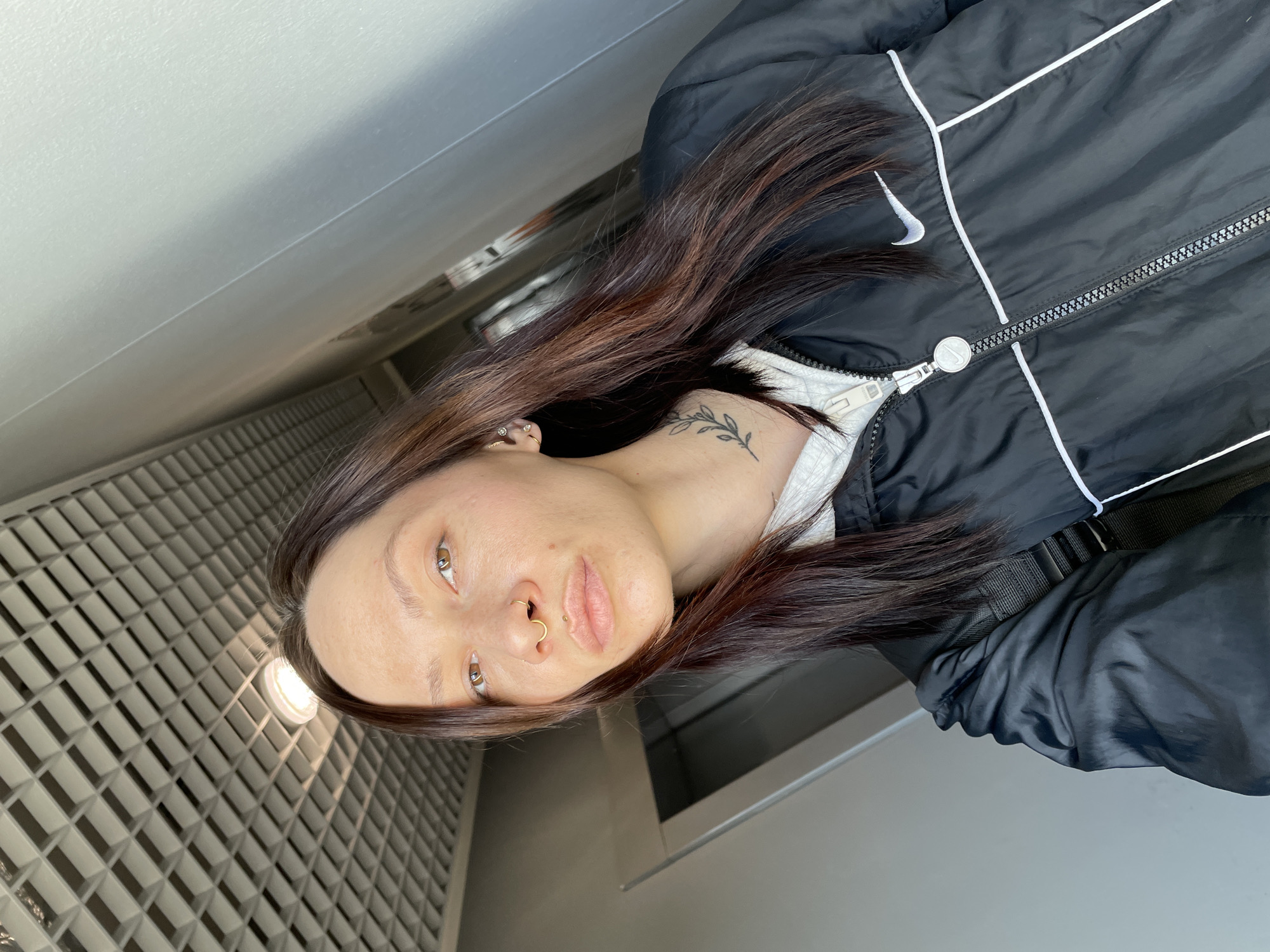 A medium shot depicts a woman wearing a black Nike jacket with white trim. She has dark brown hair with lighter brown highlights. She has a nose ring and a small tattoo on her neck. The background features a grid-like tile pattern, suggesting a public or commercial space. The setting could be an indoor location, such as a corridor or a restroom. There is no indication that this image was taken in Izhevsk, Russia.