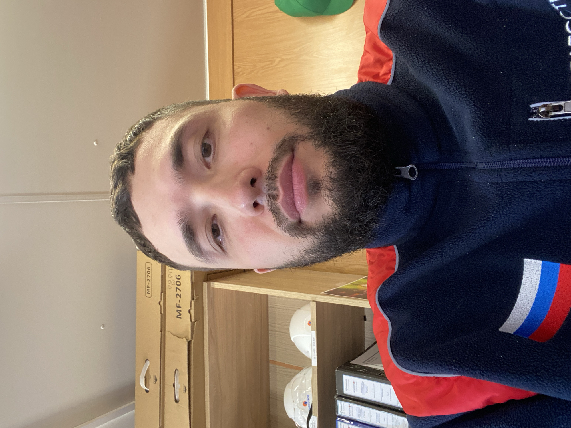 A man with a dark beard and short dark hair is shown in a close-up, head-and-shoulders shot. He appears to be inside a building or workspace, standing in front of shelves with cardboard boxes labeled "MF-2706" and some white hard hats. He is wearing a navy blue fleece jacket with red accents and a patch featuring white, blue, and red stripes reminiscent of the Russian flag. No specific location in Luzhitsy, Russia, can be identified from the image.