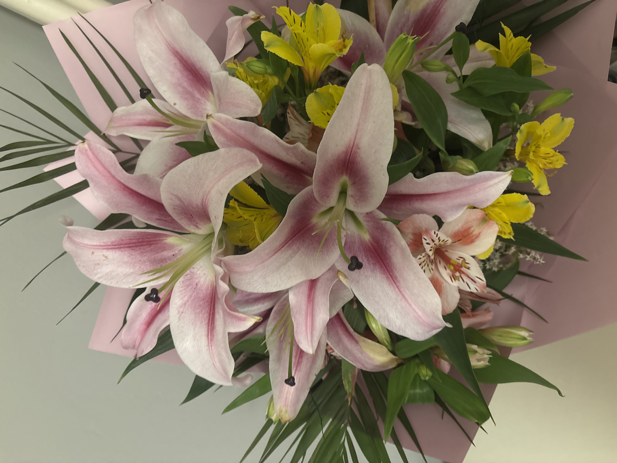 The image shows a bouquet of flowers, primarily pink and white lilies with yellow alstroemeria, wrapped in pink paper. Green foliage is interspersed throughout the bouquet. The background is a plain, light gray surface. The location is not discernible from the image. There are no people present.