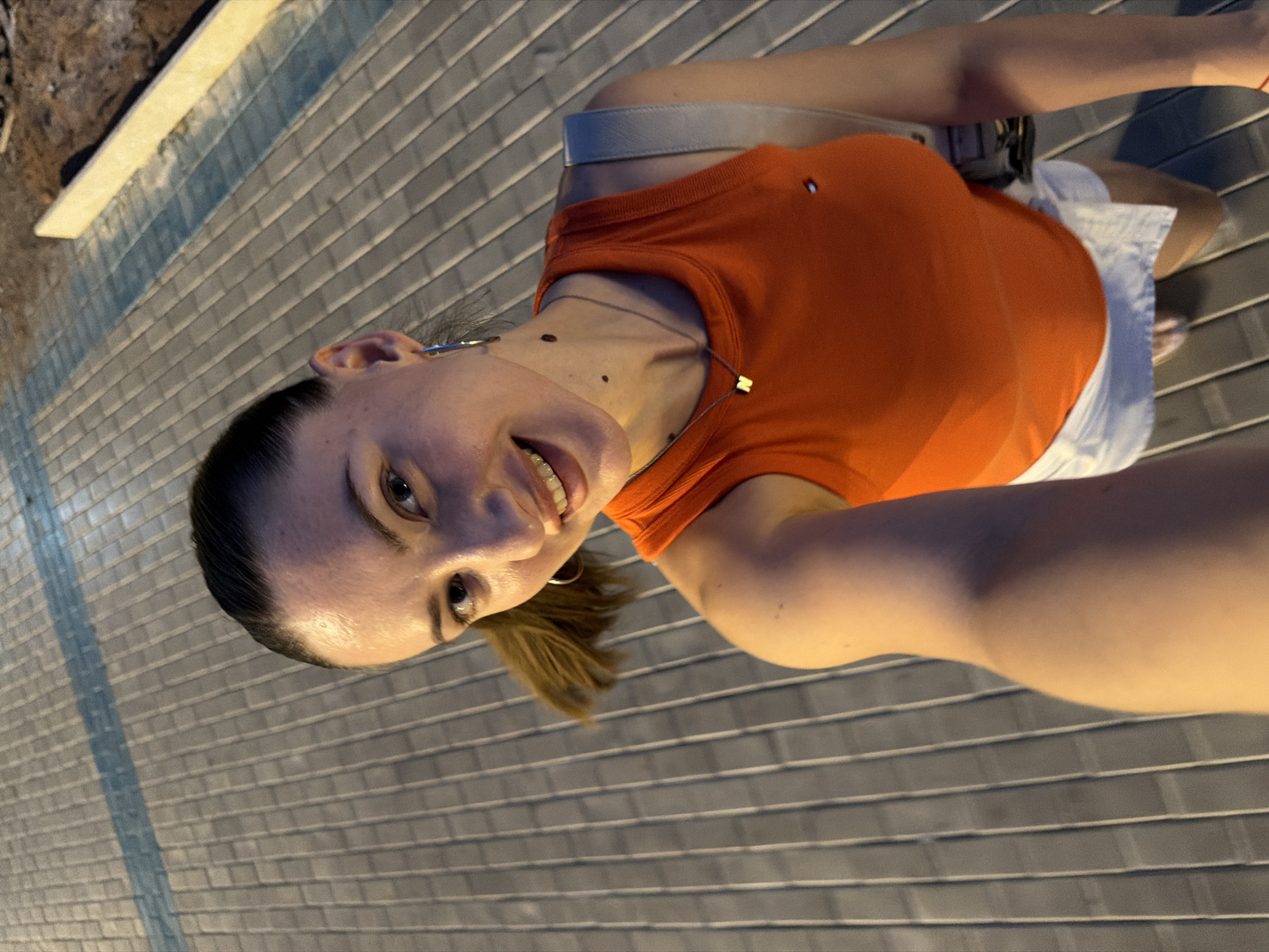 A woman with brown hair pulled back in a ponytail takes a selfie outdoors at night. She is wearing an orange tank top and a white skirt. She has a necklace with a gold "N" pendant and hoop earrings. The ground is paved with gray rectangular stones and there is a blue stripe running diagonally across the pavement. In the background, there is a construction site with dirt and a concrete barrier. The lighting suggests it is artificial, likely from streetlights or lamps. The woman is smiling at the camera.