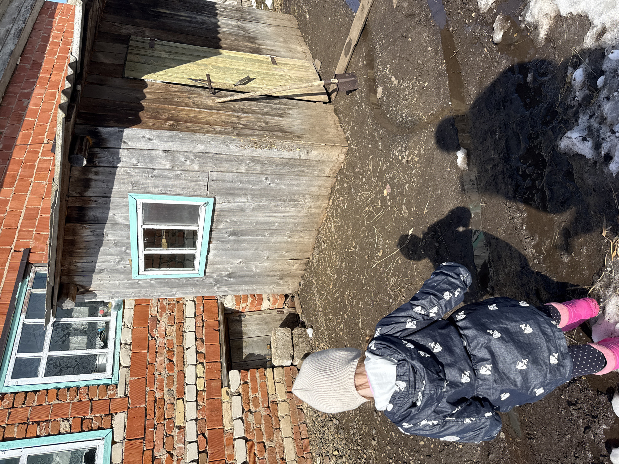 The image shows a young child standing in a muddy area near a brick and wood building. The child is wearing a patterned blue coat, a light-colored knit hat, polka dot leggings, and pink boots. The ground is a mix of mud, water puddles, and patches of snow.
The building has a section with exposed red bricks, some areas of which are deteriorating, with window frames painted light blue. Adjacent to the brick part is a wooden structure with a window and what appears to be a barred-up opening. A shovel leans against the wooden wall.
The location is presumably Кипчак, Russia, as specified. The weather appears to be cold and wet, likely early spring, judging by the melting snow and mud.