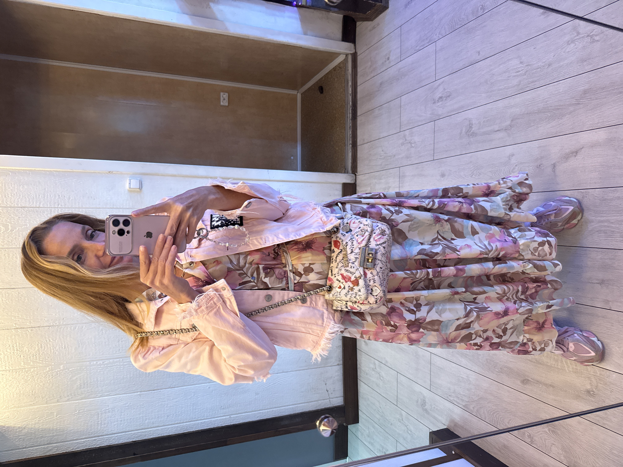 A woman is taking a mirror selfie, showcasing her outfit. She is wearing a light pink denim jacket with frayed edges and a long, flowy floral dress with pink, purple, and brown blossoms. She has a small white crossbody bag with a graphic print and a silver chain strap. Her left hand is holding up her phone, a light pink iPhone, to take the picture. She has blonde hair and is looking at her reflection.
The setting appears to be indoors, possibly a changing room or a space with mirrors. The floor is made of light-colored wood planks. Behind her, there is a wooden panel wall and a shelf or cubby with a light brown interior. The lighting suggests it is daytime. There are no overt emotional expressions visible, but the pose is casual and confident. The graphic print on her bag contains text that is partially obscured but includes words like "LOVE", "LIPS", and "KISS". The location context provided is Lomonosov, Russia.