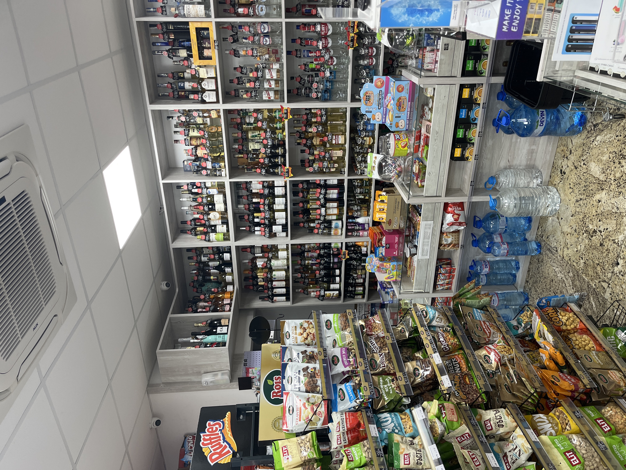 The image depicts the interior of a small shop, possibly a convenience store or liquor store, located in Sofia, Bulgaria. The scene is dominated by shelves stocked with various alcoholic beverages on the left and snacks, nuts, and water bottles on the right.
**Key features:**
* **Shelves:** Floor-to-ceiling shelves are packed with bottles of liquor and wine.
* **Snacks:** The right side of the store features angled racks of snacks, mostly nuts, and bottled water.
* **Signage:** Bulgarian Cyrillic script can be seen on product packaging and price tags. Brands like "Rois," "ELIT," "Ruffles," "Chio," and "Devin" (for water) are visible.
* **Environment:** The store has a tiled floor and fluorescent lighting. An air conditioning unit is mounted on the ceiling.
* **Security:** Security cameras are installed on the wall.