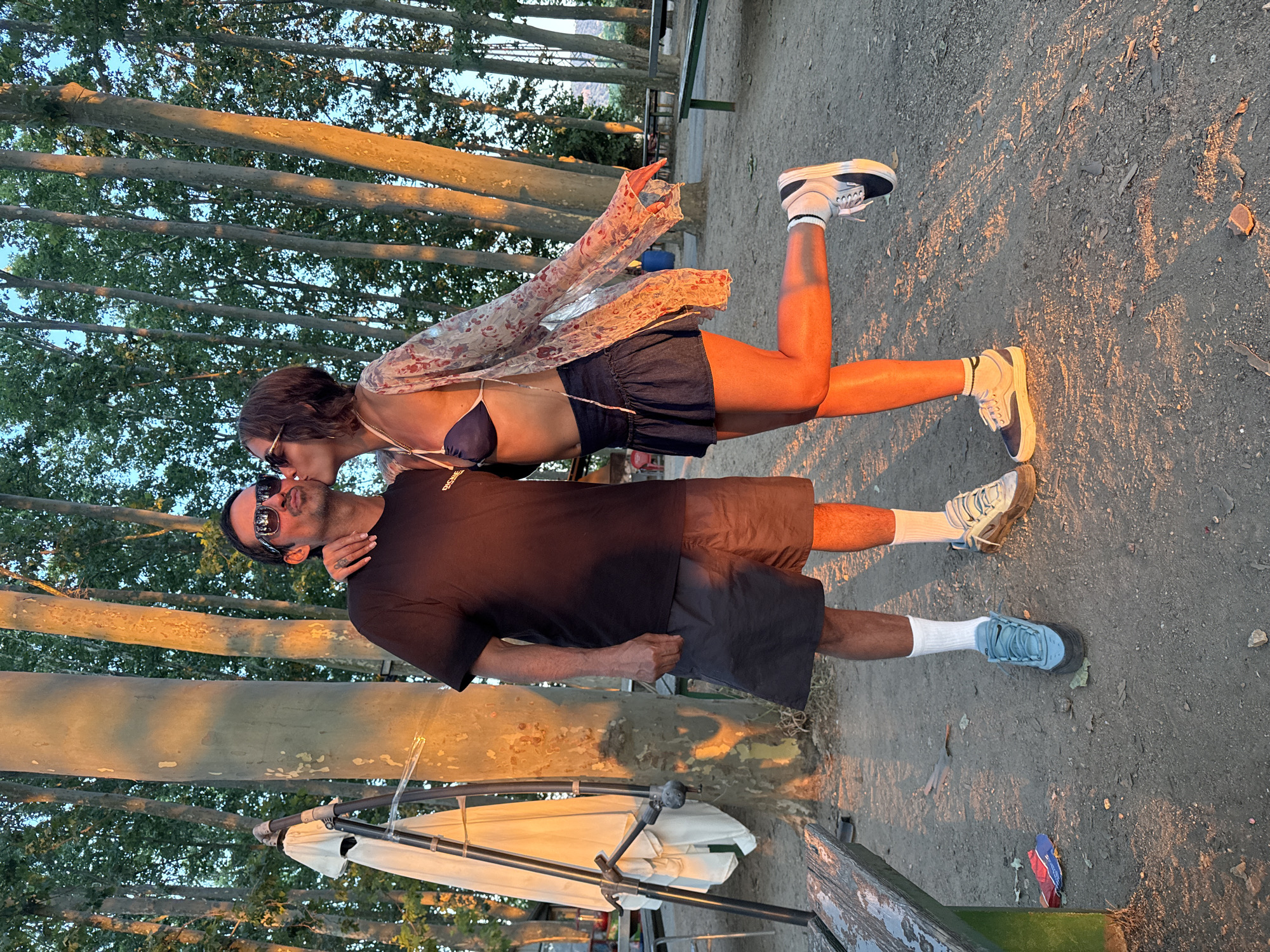 This image captures a couple in an intimate moment, with the woman planting a kiss on the man's cheek. They are standing outdoors, with a backdrop of tall trees and a clear sky. The golden hour light casts a warm glow on the scene, illuminating the dust on the ground and highlighting the textures of the tree trunks. The man, wearing a black t-shirt and dark shorts, has his eyes closed and is wearing sunglasses. The woman, in a blue bikini top, denim skirt, and a sheer, floral-patterned cover-up, is balancing on one leg with her arm around the man. In the background, there are picnic tables and benches, suggesting a park or recreational area. The presence of a ladder with a white cloth draped over it, and a discarded packet of chips near a bench, add to the informal, lived-in atmosphere of the location in Santa Cristina d'Aro, Spain.