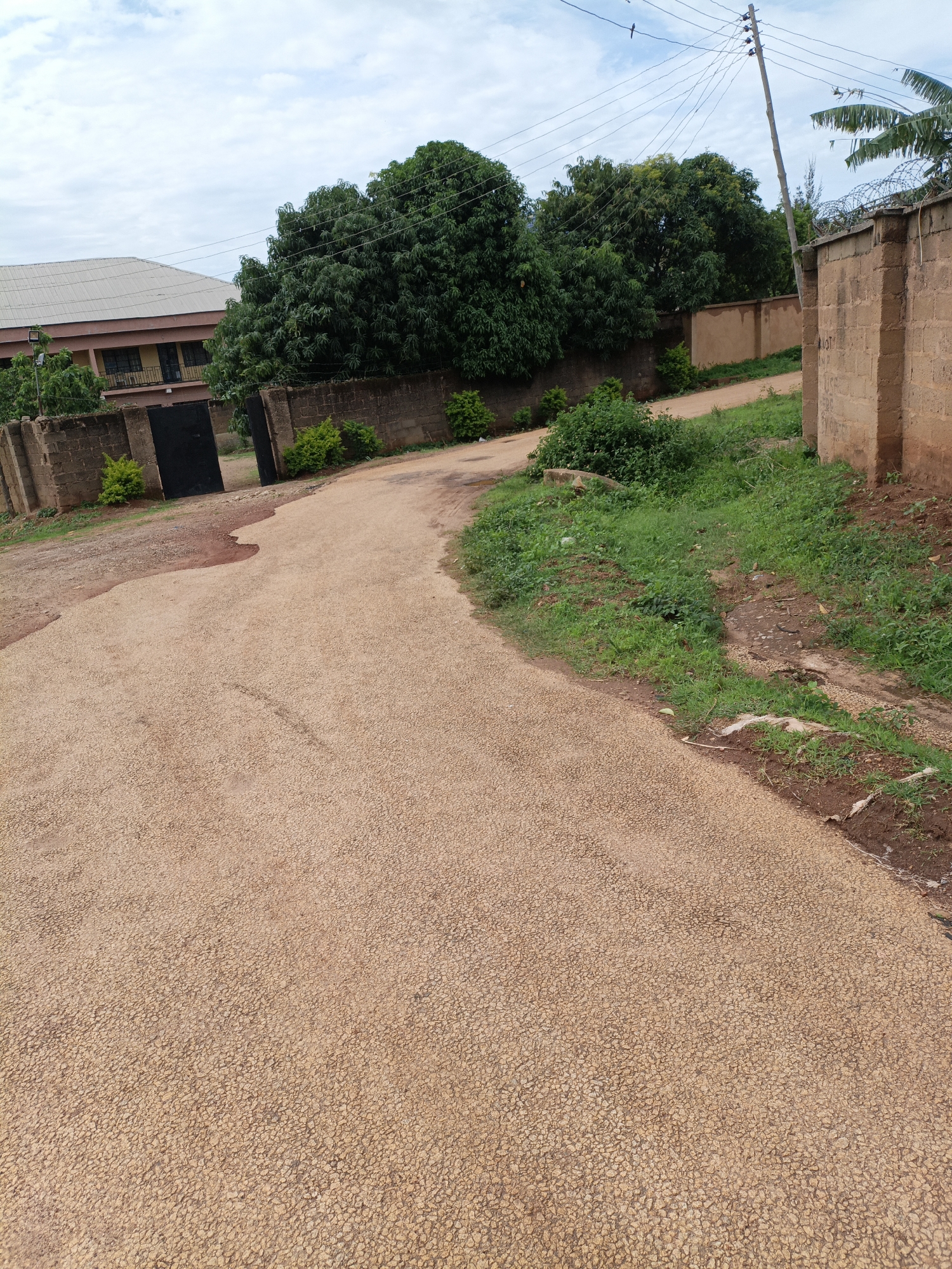 The image depicts a rural road in New Stadium, Nigeria, characterized by its light brown, pebbled surface. The road curves to the right, leading towards a large, open black gate. To the left of the gate, a two-story building with a sloped grey roof and brown walls is visible. Lush green trees and bushes are abundant throughout the scene, with some particularly large trees framing the gate and extending further into the background. A brick wall, topped with barbed wire, runs along the right side of the road, partially obscuring the view beyond. A utility pole with multiple wires stands to the right of the road, indicating the presence of electricity infrastructure. The sky is overcast with a mix of white and grey clouds. Tire tracks are discernible on the road surface.