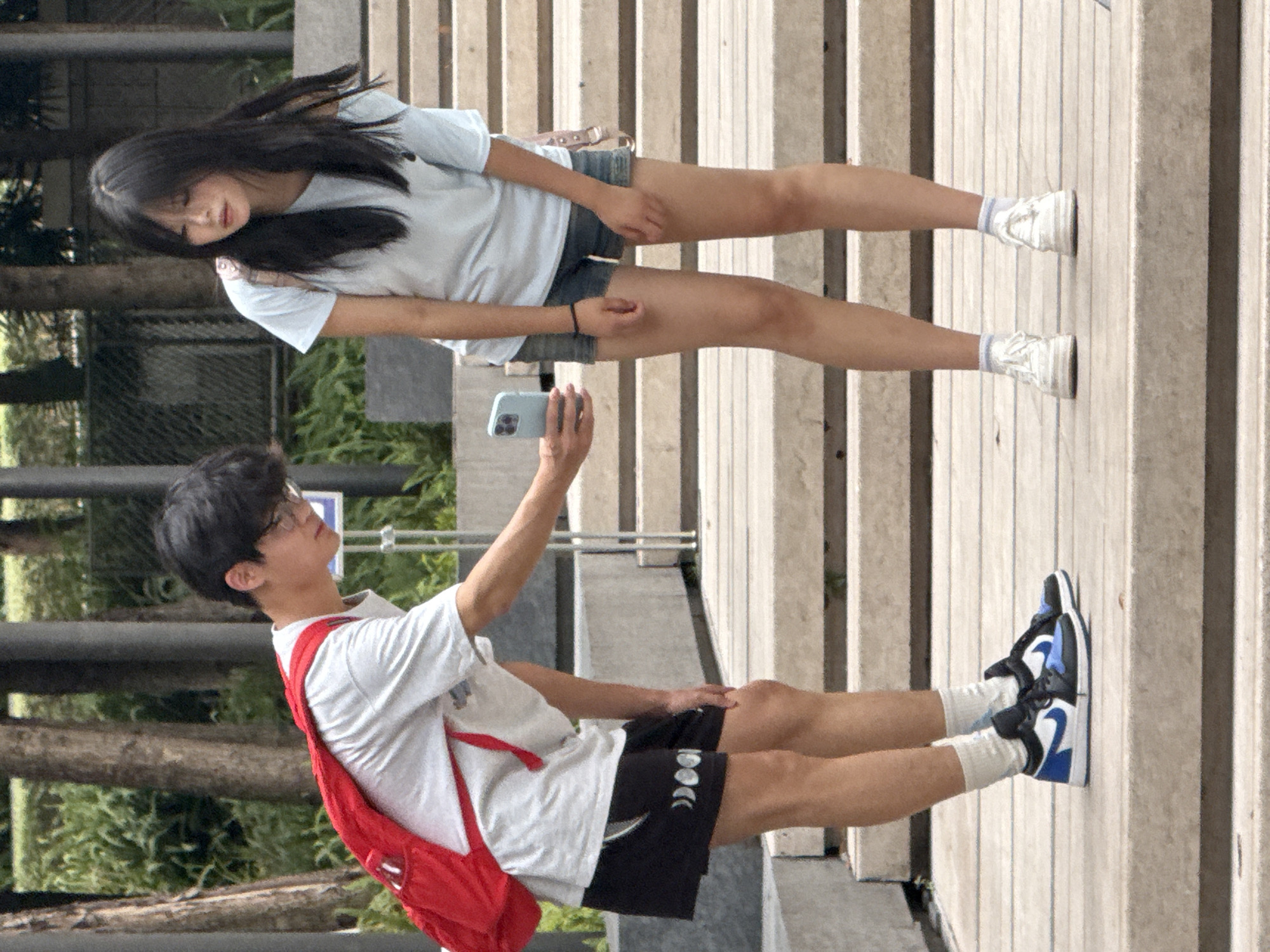 In Yuzhong District, China, two young people are standing on a tiered wooden and concrete outdoor staircase. On the left, a male wearing glasses and a white t-shirt with a red backpack is holding up a light blue smartphone. He is looking at the phone as if taking a picture or video. He is also wearing black shorts with white accents. On the right, a female with long dark hair is wearing a light blue t-shirt and denim shorts, and she is looking down towards the male. She has a light-colored backpack over her shoulders. The background consists of trees and some urban structures. The lighting suggests it is daytime.