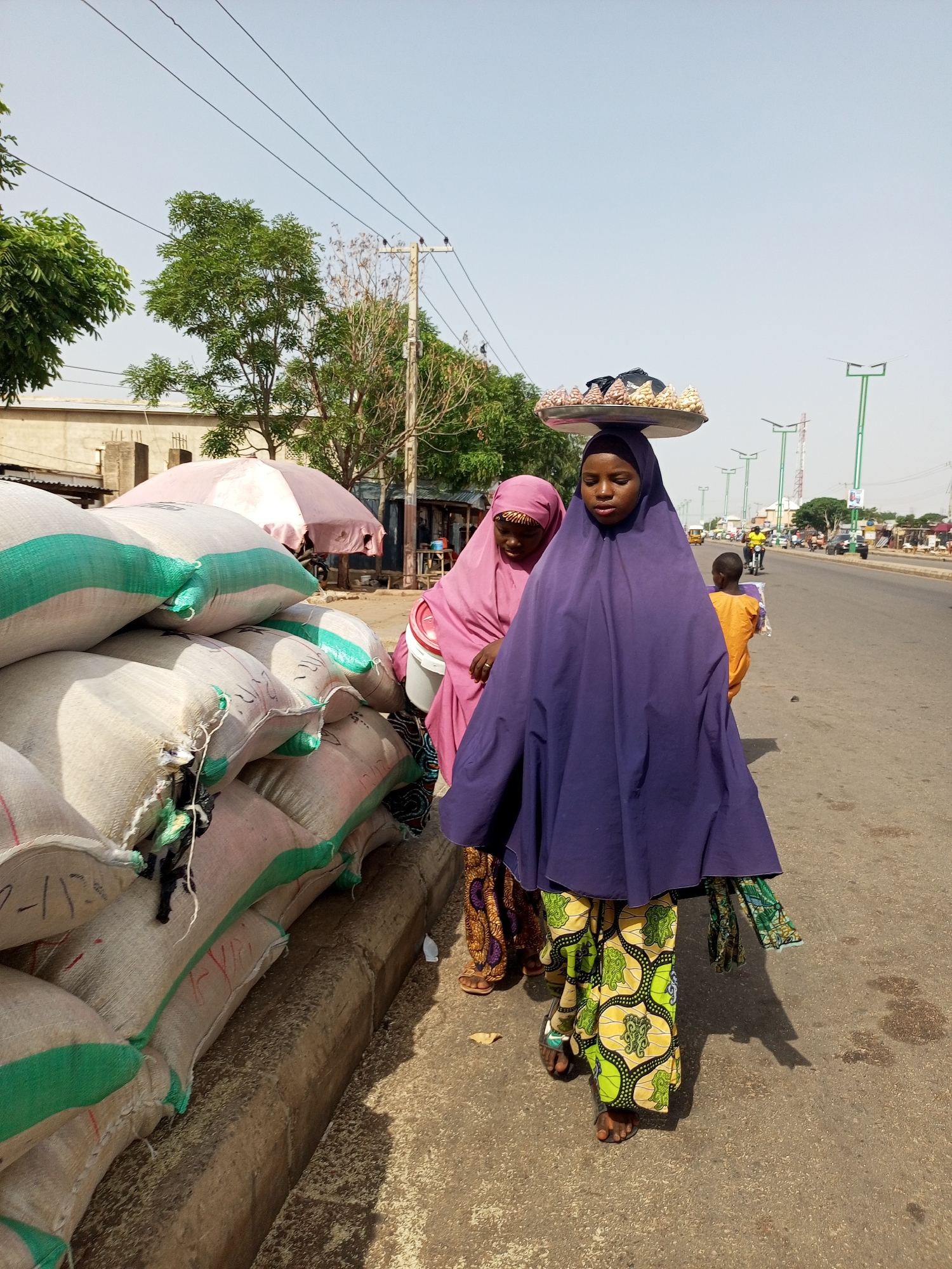 On a sunny day in Gombe, Nigeria, two women wearing headscarves and patterned skirts stand on the side of a busy road, near a pile of large sacks. The woman in front wears a violet headscarf and carries a tray of food on her head. A child in an orange shirt walks on the other side of the woman. Cars, motorcycles, and pedestrians are seen on the road in the background. Overhead power lines and solar street lights are visible above the road.