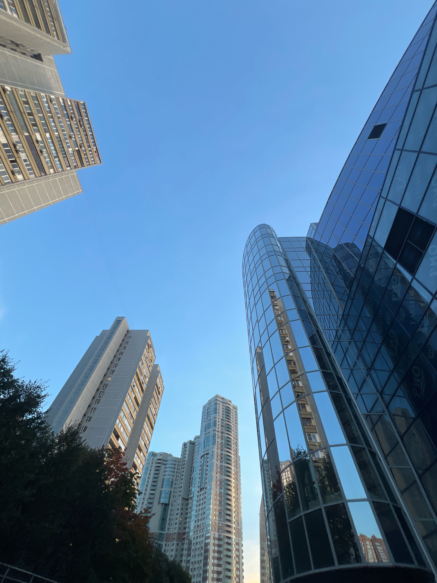 In Moscow, Russia, this image captures an upward-looking perspective of contemporary high-rise buildings against a clear blue sky. On the right, a modern skyscraper with a glass facade reflects the surrounding architecture and sky. To its left, another building features a curved glass exterior, also reflecting the sky and other structures. In the mid-ground, several residential buildings with numerous windows are visible, some appearing older and more utilitarian, while others are sleeker. A line of trees in the foreground on the left partially obscures some of the buildings. The overall scene is dominated by the geometric shapes of the architecture and the vast expanse of the sky, suggesting an urban environment.