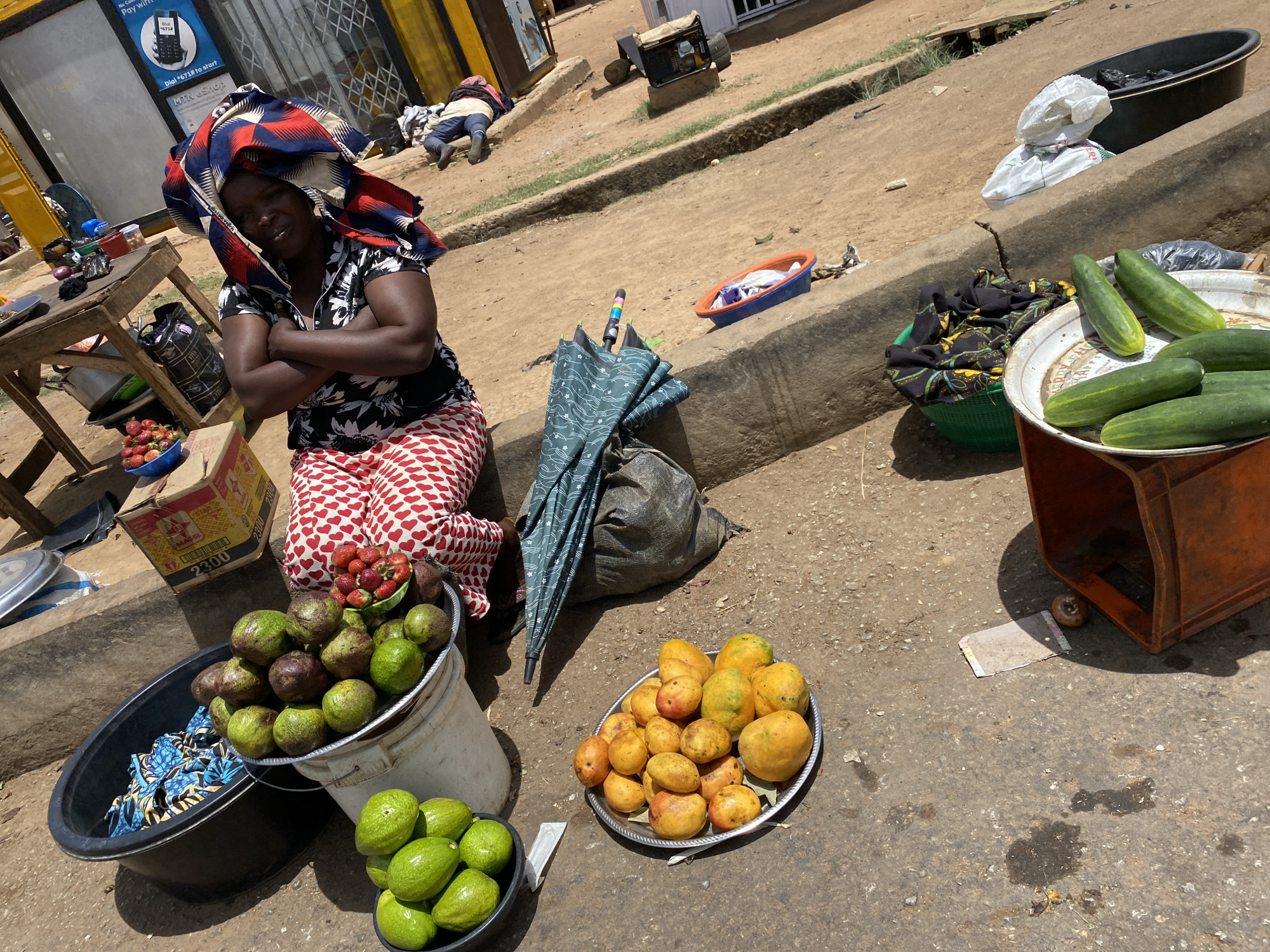 The image depicts a woman in Bukuru, Nigeria, seated on the ground beside her fruit stand. She wears a patterned headscarf, a floral blouse, and red and white patterned pants, and her arms are crossed. Her merchandise includes avocados, mangoes, strawberries, and cucumbers, displayed in buckets and on trays. An umbrella and a bag rest beside her. In the background, a person lies on the ground near a generator, and signage on a building is visible, including "MTN GSHOP."