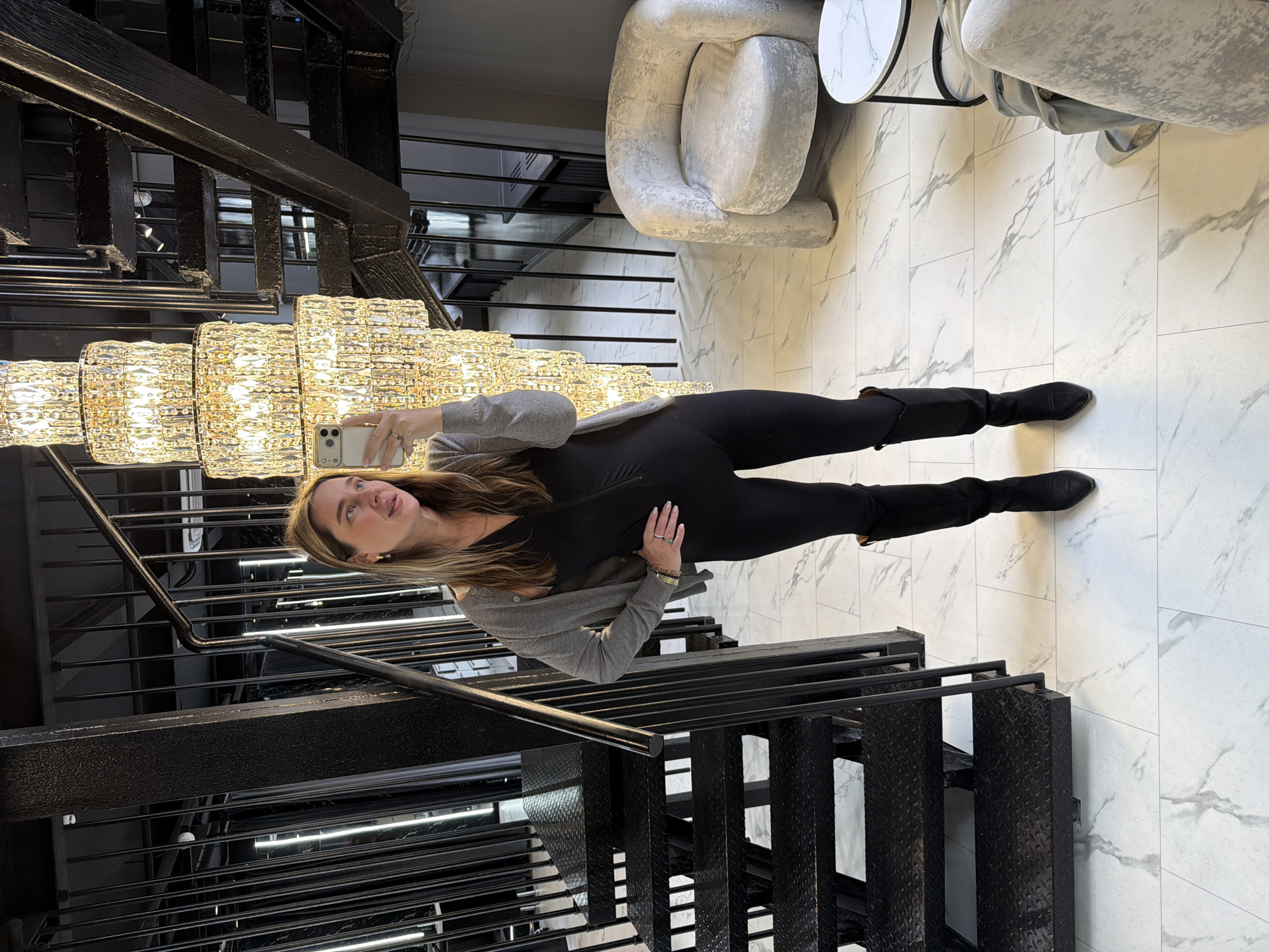 A woman stands on a white marble-patterned floor facing a large, tiered chandelier affixed to a black metal staircase. She is wearing black leggings, a black top, and a gray cardigan, and holds a smartphone in her left hand, presumably to take a photo of the chandelier. Her right hand rests on her stomach. The staircase extends behind her and above the chandelier. In the background, a round white table and a light gray, uniquely-shaped chair are visible. The specific location in Moscow, Russia is not discernible from the visual data.
