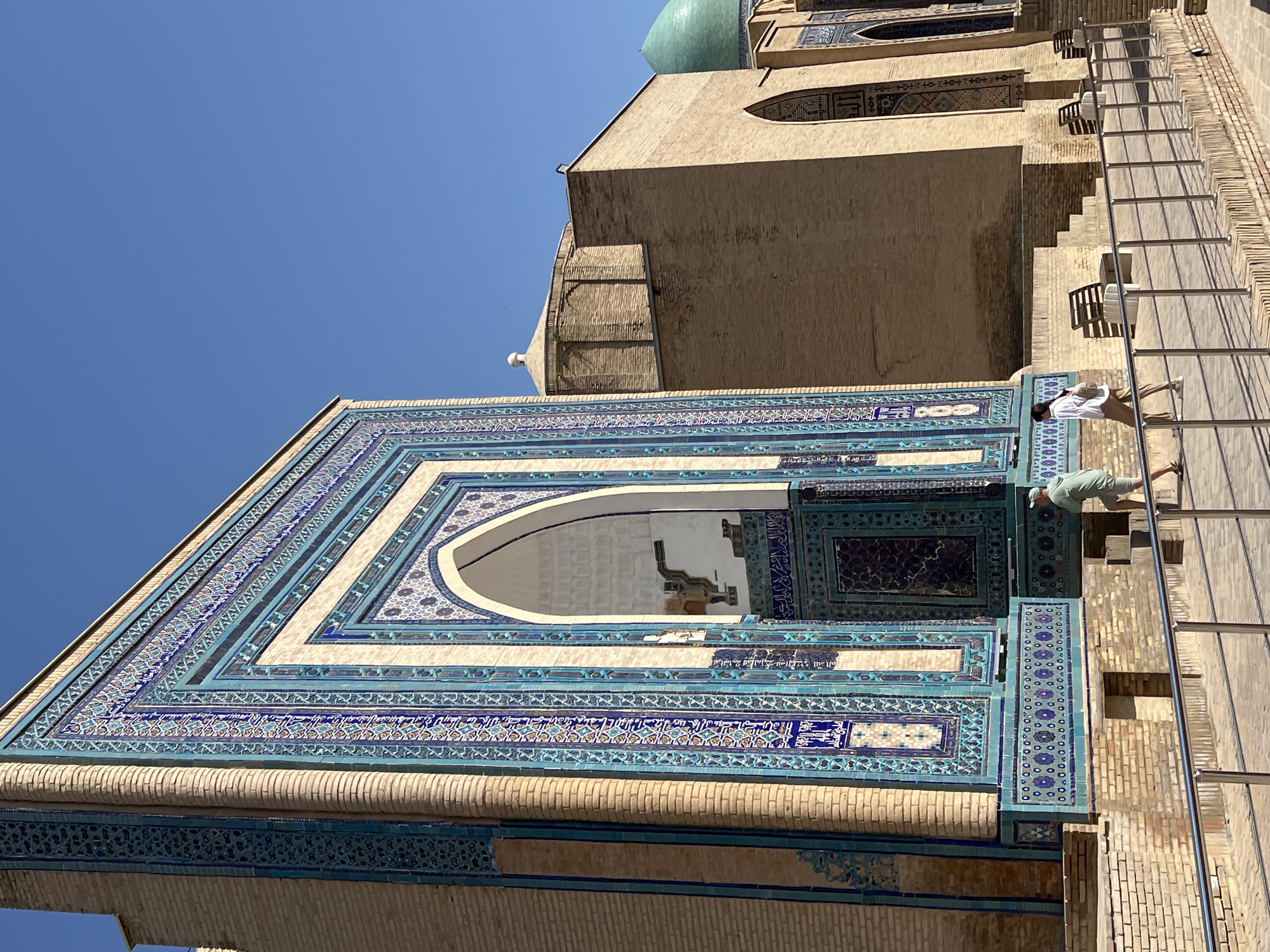 The image depicts the exterior of the Gur-e-Amir Mausoleum in Samarkand, Uzbekistan. The mausoleum is characterized by its intricate blue and turquoise tilework adorning the facade, particularly around the grand arched entrance. The brickwork of the structure is visible, contrasting with the detailed tile mosaics. In the foreground, a paved walkway is bordered by a metal railing, with several people walking and standing near benches. The sky is a clear, bright blue. Adjacent to the main mausoleum, another part of the complex, likely the mosque, is visible with a dome and brick construction. The architectural style suggests a Timurid influence, common in Samarkand.