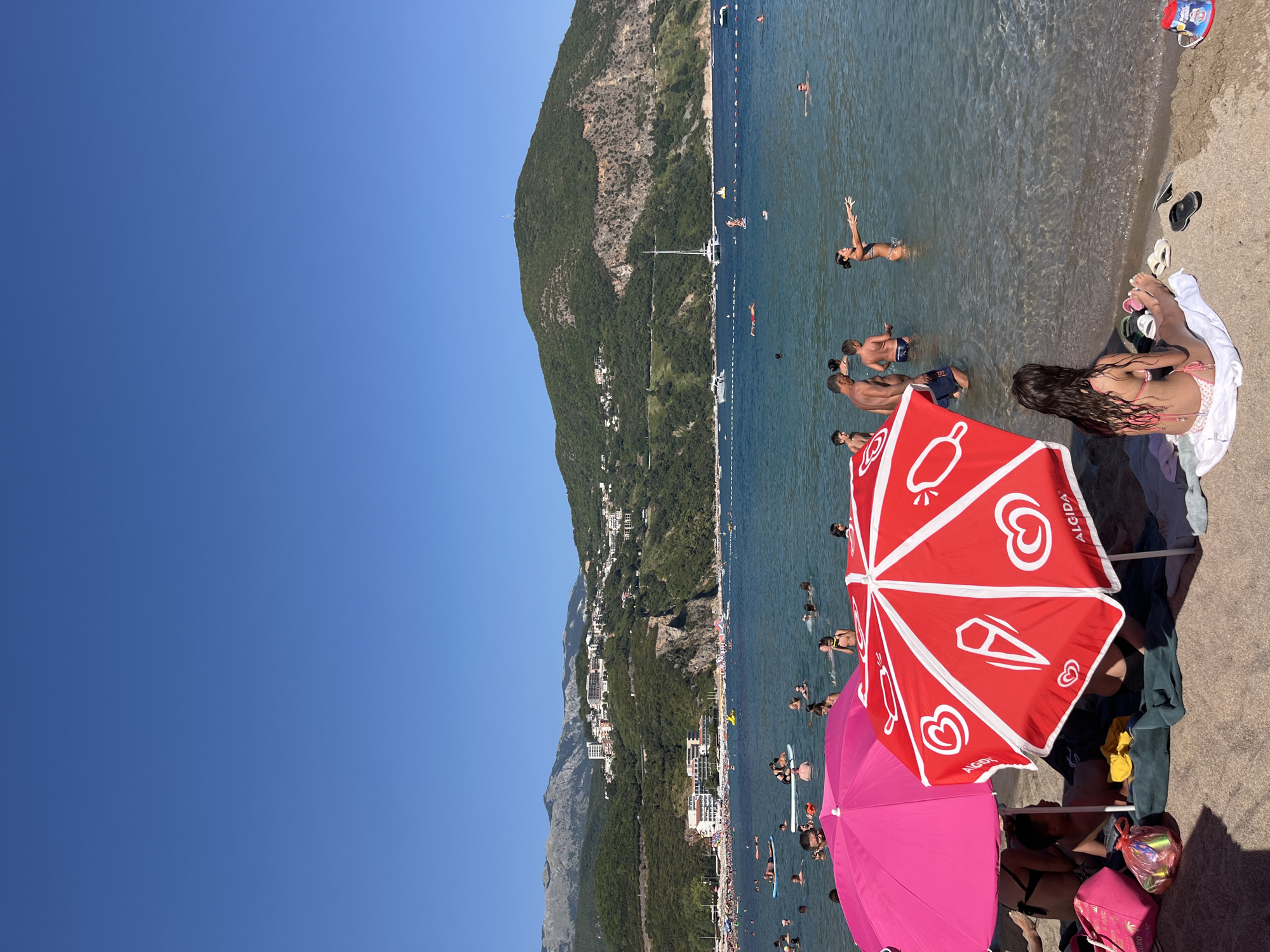 This media file depicts a sun-drenched beach scene in Prijevor, Montenegro. The main subjects are numerous people enjoying a day at the seaside. The setting is a sandy beach with clear, calm water.
The activity is recreational; people are swimming, wading in the shallow water, and relaxing on the beach. Some individuals are visible in the water, with others lounging on the sand. The presence of beach umbrellas, including a prominent red one with an Algida logo and a pink one, suggests a typical beach day.
Notable details include the bright, sunny weather, indicated by the clear blue sky and the strong sunlight casting shadows. The time of day appears to be midday or early afternoon, given the intensity of the sun. A large, green, mountainous hillside with scattered buildings dominates the background, providing a strong sense of place. The sea is populated with swimmers and some small boats or buoys marking swimming areas.
Visible text includes "ALGIDA" printed on the red umbrella. The overall mood conveyed is one of leisure and enjoyment, typical of a summer vacation at the beach.