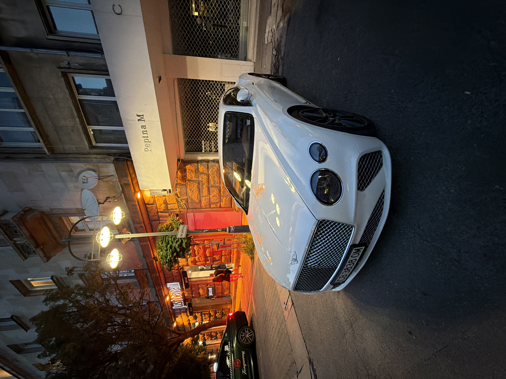 This image shows a street scene in Sofia, Bulgaria, at night. A white Bentley Continental GT is parked in the foreground, its front grille and headlights illuminated. To the left of the Bentley, a black taxi with "Tanqueray" branding and a taxi number "012" is parked. Behind the vehicles, a building facade features a lit store with the "Just Cavalli" logo. Above the store, a sign reads "Pepina M". A lamppost with three illuminated globes stands on the sidewalk. The overall lighting suggests it is dusk or nighttime.