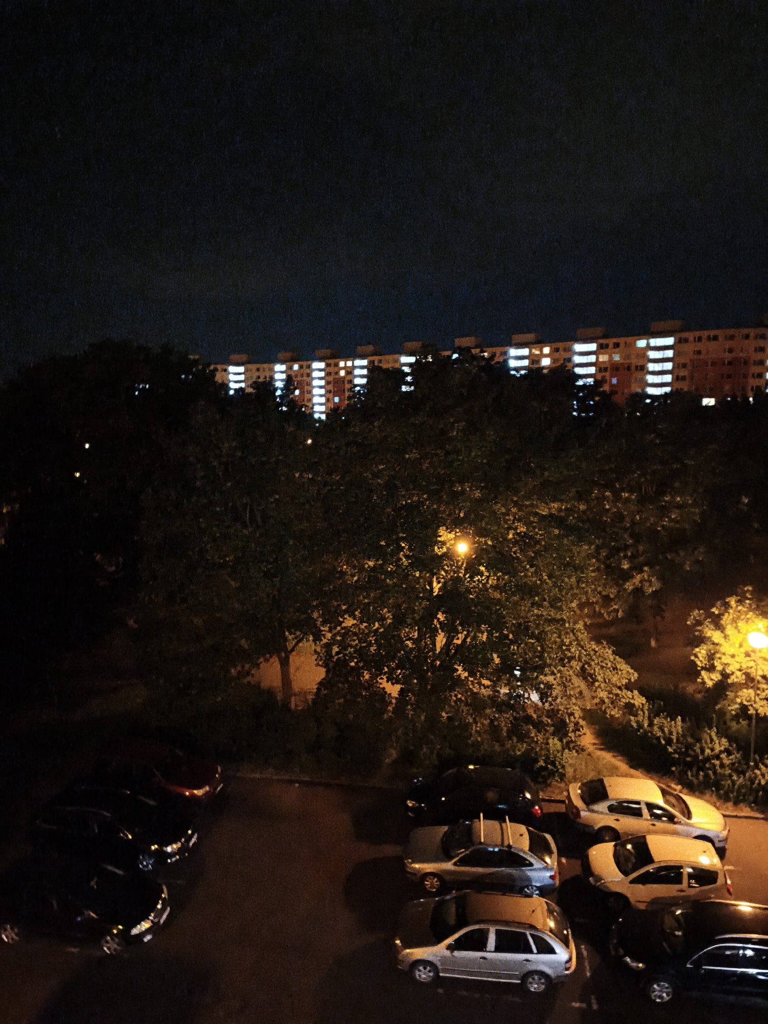 This is a night-time, high-angle shot of a residential area in the city of Most, Czechia. The scene shows a row of apartment buildings in the background, with many windows lit up, indicating occupancy. In the foreground, a parking lot is visible with several cars parked. Trees are scattered throughout the scene, some with foliage illuminated by streetlights. The overall impression is a typical urban night scene with evidence of human activity in the buildings and the presence of vehicles.