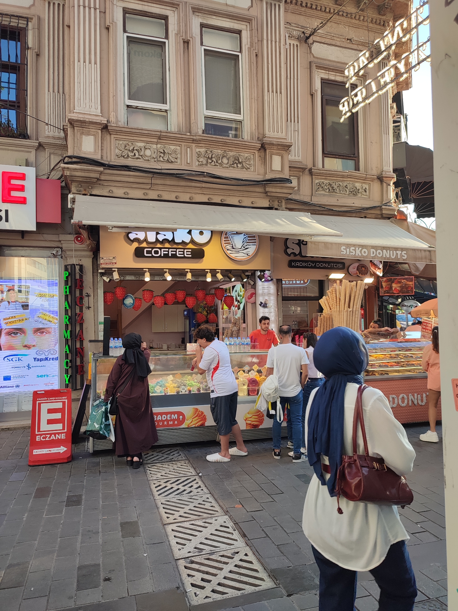 In Kadıköy, Turkey, a street scene unfolds with multiple businesses and pedestrians. On the left, a brightly lit sign displays large red letters "E" and "ECZANE" indicating a pharmacy. Next to it, a digital billboard shows faces and text in Turkish. A prominent awning stretches across the scene, covering a storefront selling coffee and donuts. The "SisKo Coffee" and "SisKo DONUTS" signs are clearly visible. In front of the donut shop, an employee in an orange shirt serves customers from behind a display case filled with colorful ice cream. A stack of waffle cones stands prominently near the entrance. Several people are browsing or being served. To the right, a woman in a blue hijab walks away from the camera, carrying a burgundy handbag. The street is paved with cobblestones, and drainage grates are visible. The architecture of the buildings above the shops features ornate details and multiple windows. The overall atmosphere suggests a busy urban commercial area on a sunny day.