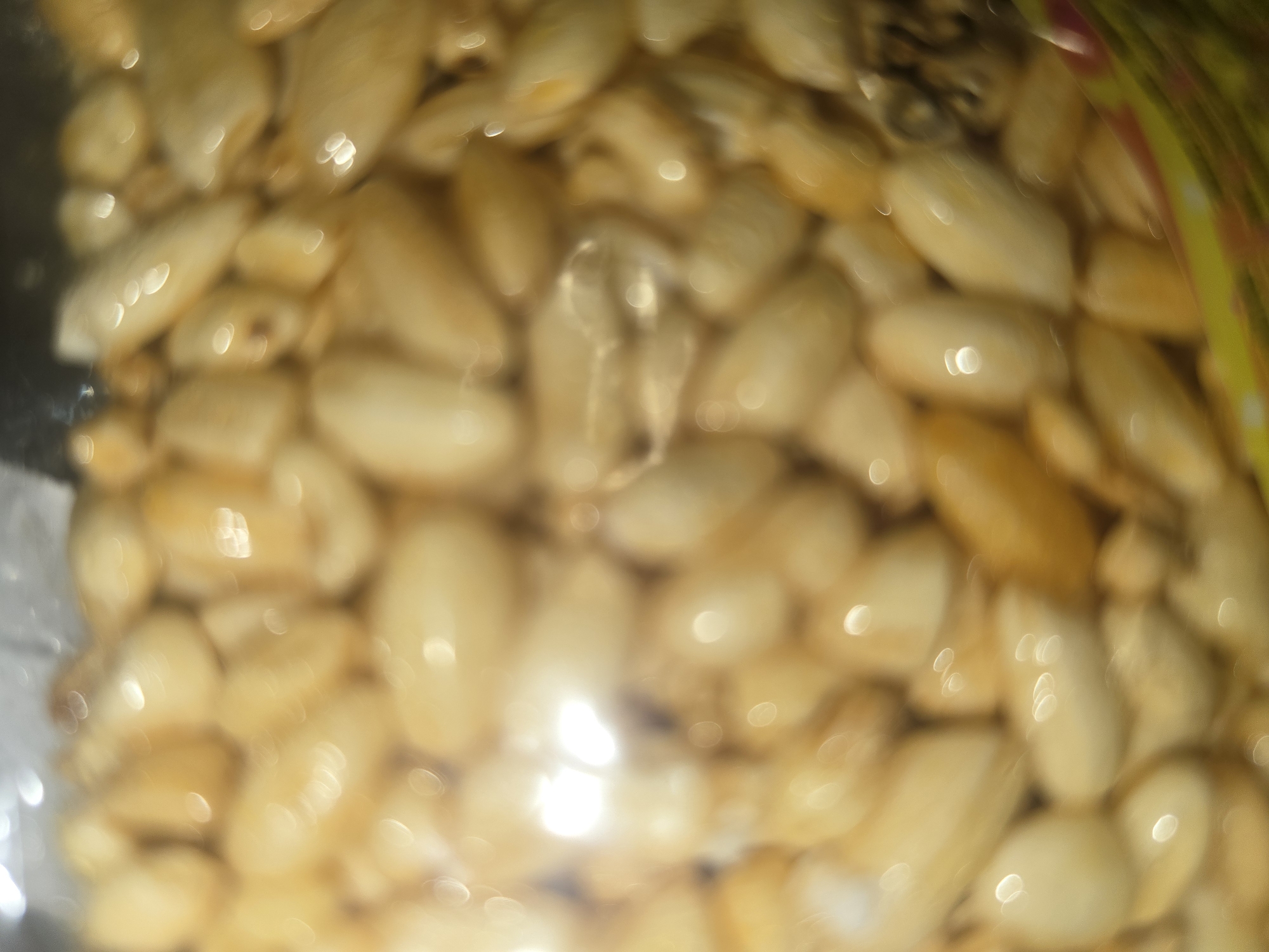 The image displays a close-up, blurry view of numerous light-yellow, oval-shaped objects, possibly nuts or seeds, contained within clear packaging. The lighting creates bright, circular reflections on the surfaces of these objects, suggesting they might be polished or coated. The background is dark and indistinct, with some hints of color in the lower right corner, potentially indicating a label or wrapper. There are no discernible human figures, actions, or interactions in the frame. The image's focus is solely on the texture and arrangement of the contained items. The location in Ulan-Ude, Russia is not visually represented in the image.