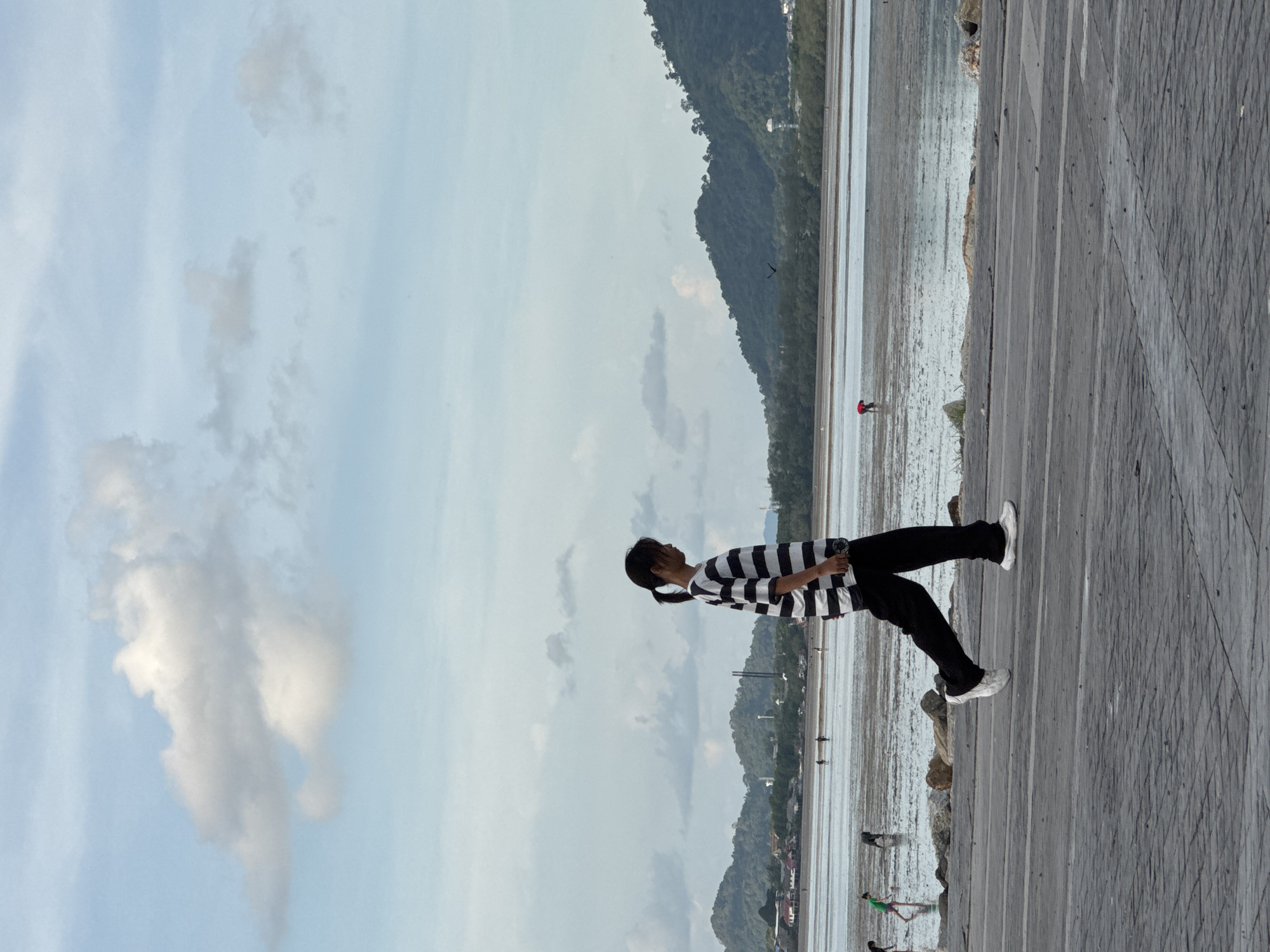 A young woman with dark hair pulled back in a ponytail is walking from left to right in the foreground of the image, her body angled away from the viewer. She is wearing a black and white striped t-shirt and black pants, with white sneakers. Her left leg is extended forward, mid-stride, and her right arm is slightly bent at the elbow.
The background reveals a wide, muddy expanse of a beach during low tide, with numerous small figures scattered across it, some appearing to be wading or collecting something from the mud. In the distance, a line of lush green, tree-covered hills forms the horizon. A calm body of water separates the foreground from the hills. The sky above is a pale blue, with scattered white clouds, some fluffy and large, others wispy and thin. The overall scene suggests a coastal location in Phuket City Municipality, Thailand, during either early morning or late afternoon, given the soft light. The ground in the foreground is paved with gray rectangular stones, arranged in a pattern.