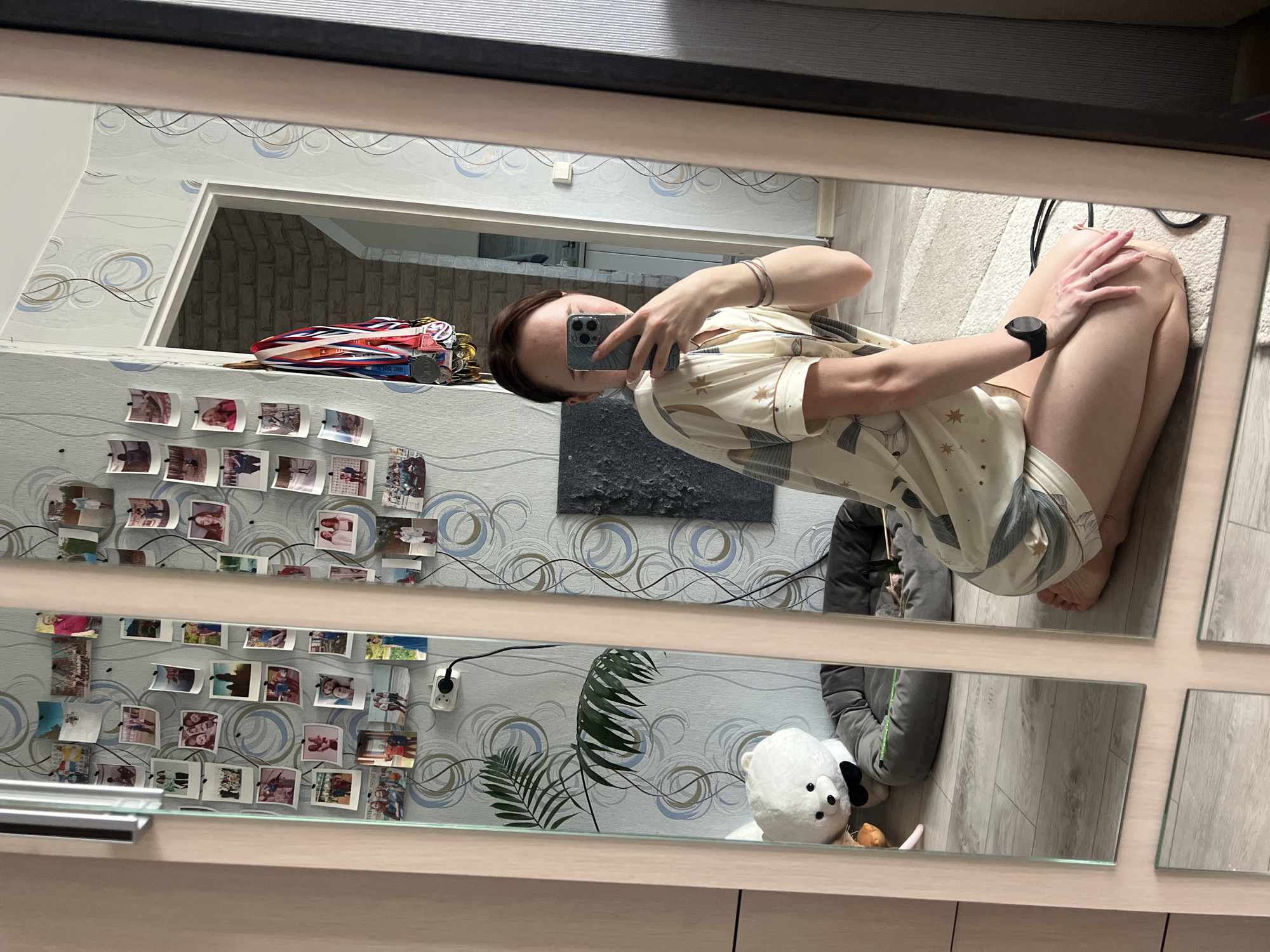 A woman is taking a selfie in a mirror comprised of multiple panels. She is wearing pajamas and a watch on her left wrist. The mirror reflects a room with light-colored wallpaper featuring circular designs, adorned with numerous small photographs arranged in rows. A ledge above the photographs holds medals and coins. A black abstract artwork hangs on the wall. A potted plant and a stuffed bear are visible near the floor. The flooring appears to be light-colored wood. The woman is holding a smartphone in her right hand to take the picture. This scene is situated in what is most likely Chaikovsky, Russia.