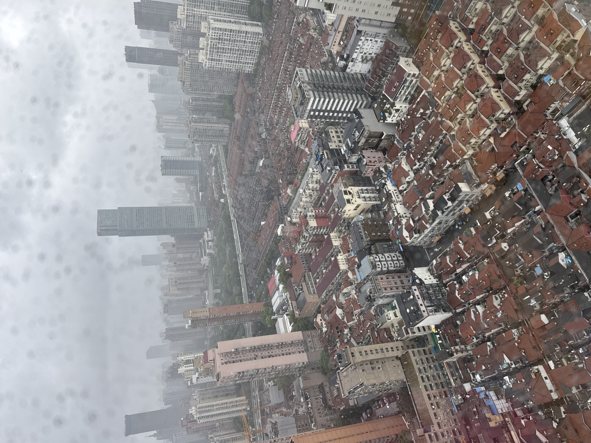 This is an aerial, high-angle photograph of a dense urban cityscape in Shanghai, China, possibly taken during or after rainfall, as evidenced by the water droplets on the camera lens. The scene is characterized by a mixture of high-rise modern buildings and older, low-rise residential structures with red tile roofs.
In the distance, several prominent skyscrapers dominate the skyline, including a very tall, slender building towards the center-right and another broad, rectangular building to its left. These modern structures contrast sharply with the tightly packed residential blocks in the foreground and midground. These older buildings are predominantly two to four stories high, with uniform red-tiled sloped roofs, indicative of a traditional Shanghai architectural style, possibly a "Shikumen" or similar neighborhood.
The streets are narrow and appear to be lined with parked cars, though the resolution and weather conditions make precise identification difficult. Some roads have visible traffic, and pedestrian activity is not discernable. The overall impression is one of a bustling city with a rich history, where old and new urban development coexist. The weather appears overcast and gloomy, with a thick haze obscuring the far distance. The presence of numerous water droplets on the lens creates a diffused, somewhat blurry effect across the entire image.