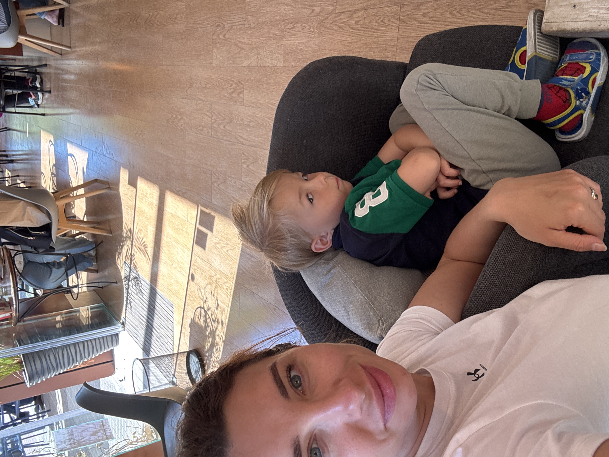 The image is a selfie-style shot featuring a woman and a young child indoors in València, Spain. The woman, positioned in the foreground, has brown hair and light eyes, offering a subtle smile to the camera. She wears a plain white t-shirt with a black "Under Armour" logo visible on the left chest. A silver ring is visible on her left hand.
Next to her, a young child, likely a boy, with blonde hair and light eyes, is resting on a dark grey armchair or couch. He is dressed in a green and navy blue polo shirt, which has a white number "3" or "B" on the sleeve, light grey sweatpants, and brightly colored blue and red Spiderman-themed sneakers with red socks. He appears relaxed, looking towards the camera or slightly off-frame.
The background reveals a brightly lit indoor environment with a warm-toned wooden floor. Numerous chairs and tables are scattered, along with glass partitions or displays and some plant pots, suggesting a public space such as a cafe, restaurant, or hotel lobby. Strong sunlight streams into the room, casting distinct, long shadows of furniture and window frames across the floor, indicating it is daytime, possibly morning or afternoon. The overall atmosphere appears casual and comfortable.