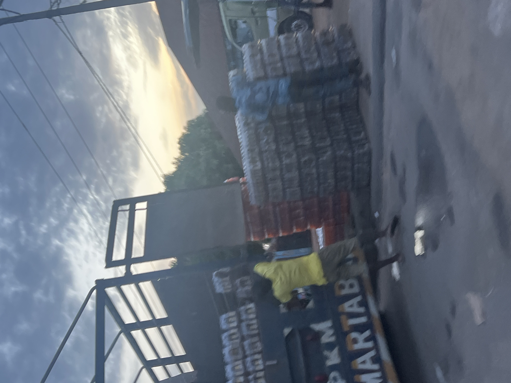 The image depicts a street scene in Jalingo, Nigeria, likely during the early morning or late evening, given the dusky sky with streaks of orange and yellow. In the foreground, a large truck is parked, its dark blue side emblazoned with white lettering that appears to read "PKM MARTAB". A person wearing a bright yellow shirt and tan pants is seen standing near the rear of the truck, appearing to be in the process of loading or unloading items.
To the right of the truck, large stacks of what appear to be bottled water are piled high. The bottles are clear and arranged in a grid pattern within translucent plastic wrapping. A person wearing a blue shirt is partially visible standing behind these stacks, interacting with the scene. Further to the right, a green truck is parked, and a portion of a building with a reddish-brown roof is visible in the background, suggesting a commercial or market area. The ground is paved and appears wet in places, with reflections of light visible. Overhead, power lines crisscross the sky, and the silhouette of trees can be seen against the horizon. The overall impression is one of commerce and logistical activity taking place in an urban environment.