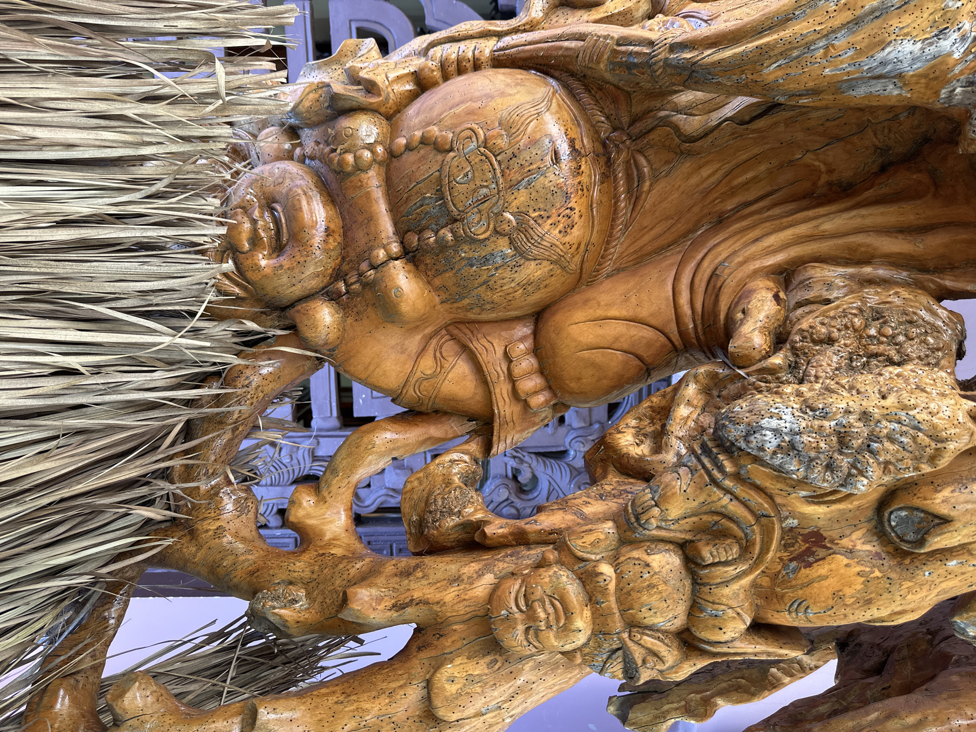 A close-up, eye-level shot captures a wooden carving of a laughing Buddha figure. The carving, made of polished wood with a warm, orange-brown hue, features intricate details such as a smiling face, a prominent belly, and draped clothing. The figure is adorned with a beaded necklace and is supported by what appears to be a carved tree trunk with a rough, textured surface. Overhead, dried straw or grass hangs down, partially obscuring the top of the carving. The background is blurred, revealing glimpses of purple and blue decorative elements, suggesting an outdoor or semi-outdoor setting, possibly in Thôn 5, Vietnam.