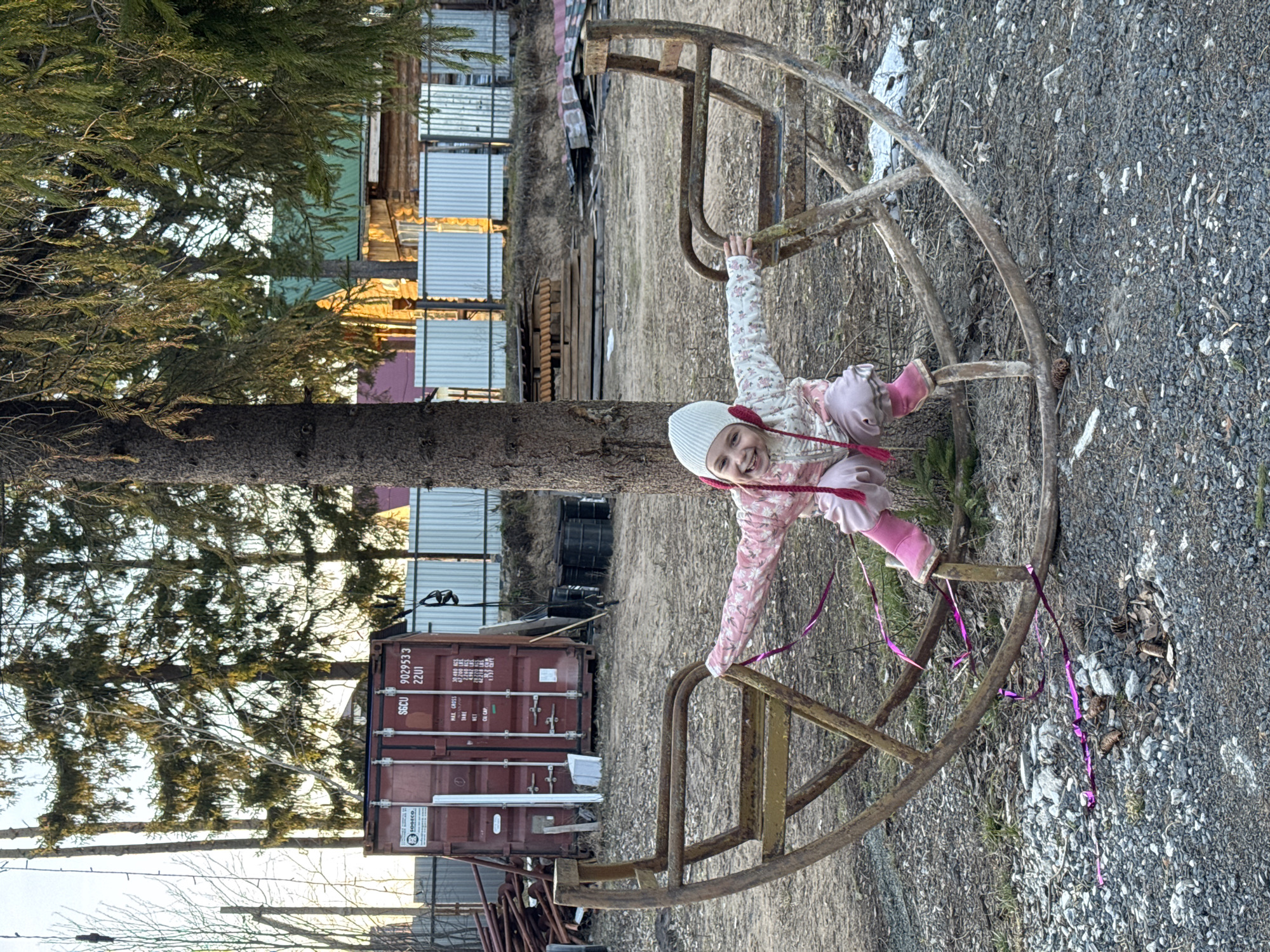 The image shows a young girl in a white hat and pink outfit playing on a rusty, old merry-go-round. The location appears to be in Chaikovsky, Russia. In the background, there is a shipping container, a wooden fence, and some trees. The ground is covered in gravel.