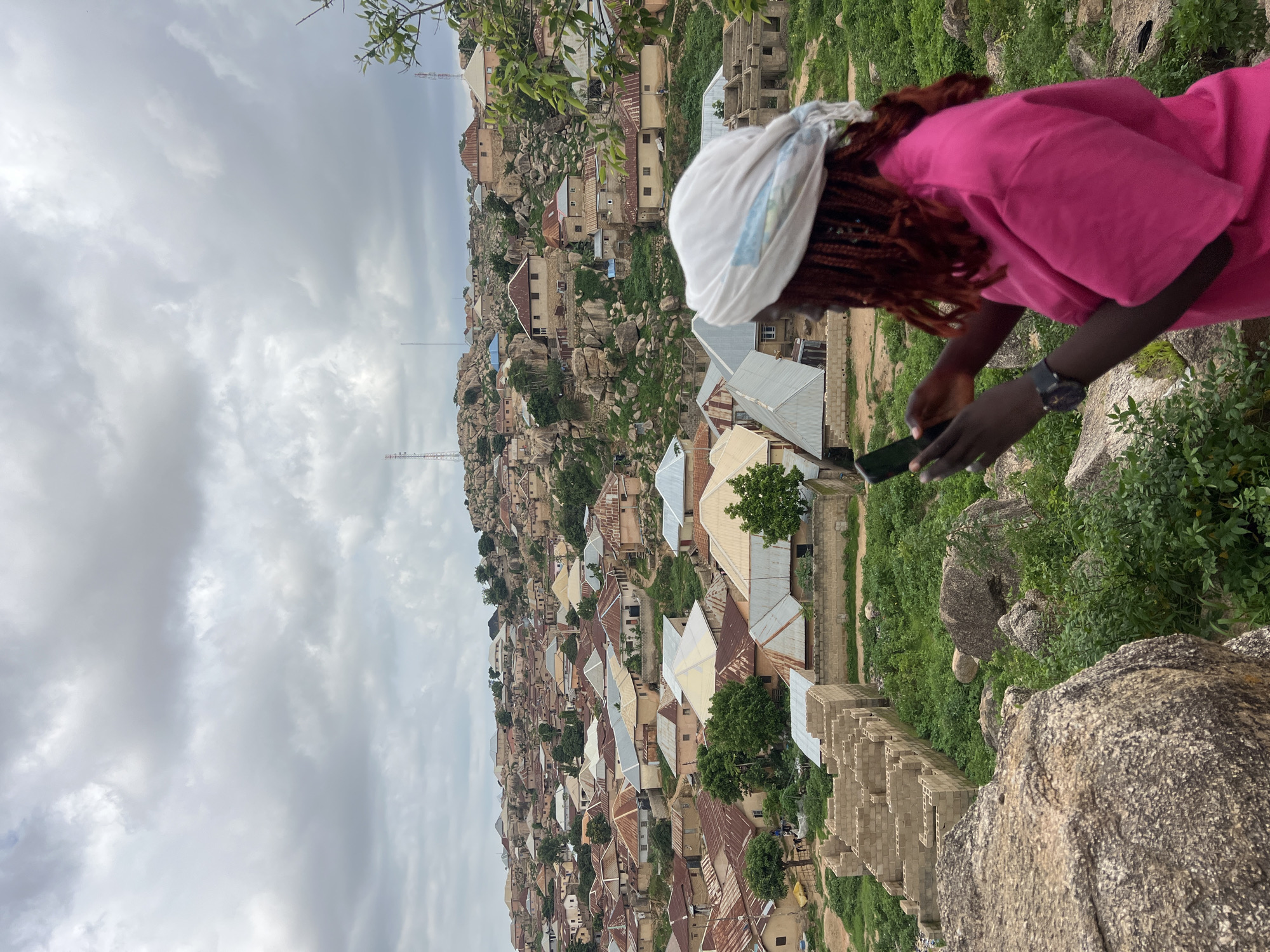 This is a medium shot of a young Black woman with braided, reddish-brown hair and a white headscarf. She is wearing a bright pink t-shirt and a black watch on her left wrist. She is holding a dark smartphone in both hands, appearing to take a picture or video. She is standing on a rocky, uneven surface covered in green vegetation, looking out over a densely populated hillside settlement.
The settlement, which appears to be located in Jos, Nigeria, is characterized by numerous buildings with corrugated metal roofs in varying shades of gray, brown, and orange, interspersed with some pale yellow structures. The houses are tightly packed together and built on a rugged, rocky terrain. Many of the buildings are constructed with mud bricks and concrete blocks. In the foreground, to the left of the woman, a partial view of a building under construction is visible, made of stacked concrete blocks. The sky above is overcast with gray clouds. Communication towers are visible on some of the higher rocky outcrops in the background. The overall impression is of a vibrant but somewhat rugged community situated in a hilly landscape.
