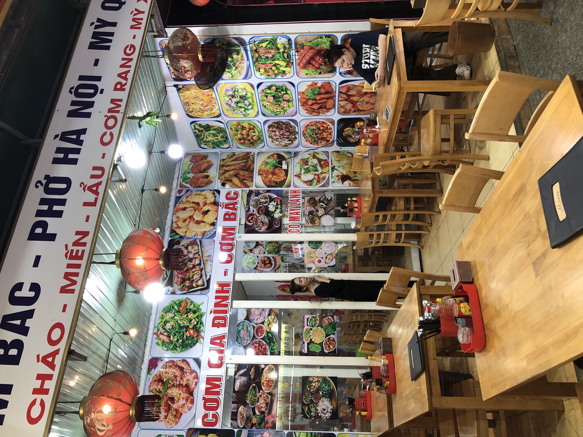 The image depicts the interior of a Vietnamese restaurant in Da Nang, Vietnam, likely specializing in Vietnamese cuisine. The restaurant features a large sign overhead with Vietnamese text, possibly indicating menu items or the restaurant's name. Red lanterns hang from the ceiling, contributing to the ambiance. The walls are adorned with a grid of framed pictures showcasing various dishes, including seafood, vegetables, and what appear to be noodle or rice-based meals. Wooden tables and chairs are arranged throughout the dining area, with some tables set with condiments in red trays. In the foreground, a wooden table is visible with a black menu. In the mid-ground, a woman in a black outfit stands near the entrance, and another woman sits at a table, using her phone. The lighting suggests it is evening or an indoor setting.