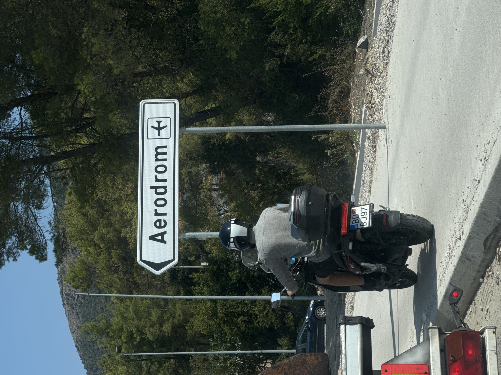 A person is riding a black scooter on a paved road. They are wearing a gray shirt and a black helmet with white accents. The scooter has a black storage box on the back and a license plate that reads "BD BJ97". To the right of the scooter, a white sign with black lettering points to the left and says "Aerodrom" with a small airplane icon. The sign is mounted on a metal pole. In the background, there are trees and a hill. A black car is parked on the left side of the road. In the foreground, part of a white vehicle with red lights is visible. The scene appears to be in Mrčevac, Montenegro, as indicated by the presence of the "Aerodrom" sign, which refers to the Tivat Airport, located in the vicinity of Mrčevac.