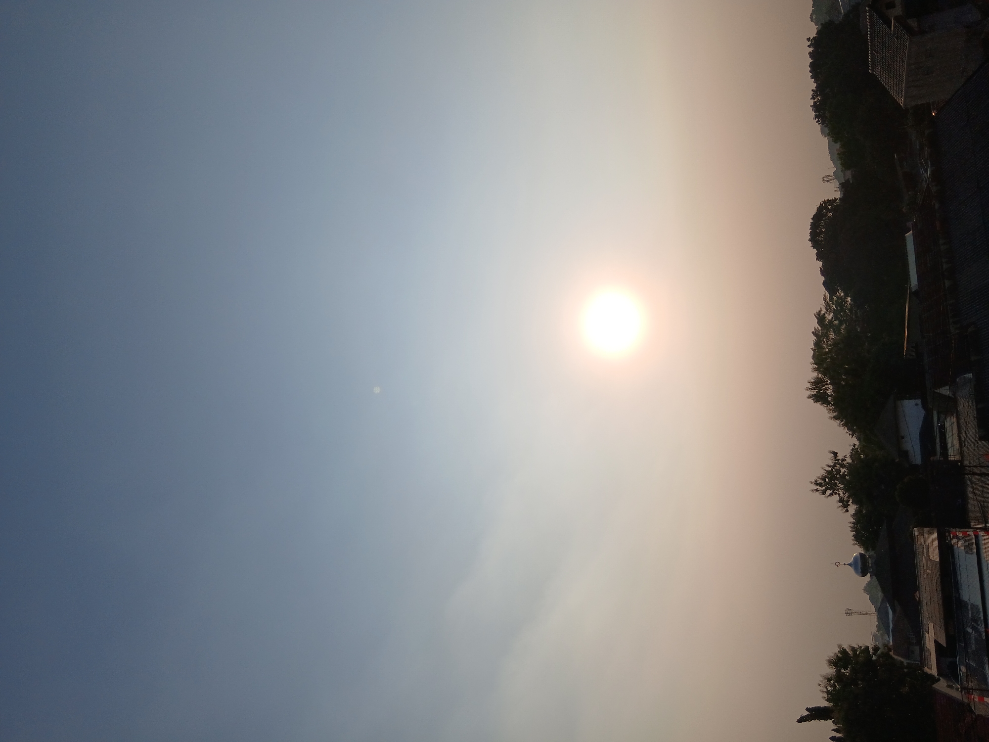 The image depicts a hazy, sunlit sky over a residential area in South Tangerang, Indonesia. The sun, a bright, luminous orb, is positioned in the middle of the frame, casting a warm glow that filters through a thin layer of haze. The sky transitions from a pale blue at the top to a lighter, almost white hue closer to the horizon.
Below the sky, a silhouetted treeline forms a dark band across the lower portion of the image. Among the trees, rooftops of houses and buildings are visible, some with corrugated metal roofs. A prominent feature is a building with a silver dome, likely a mosque, topped with a crescent moon finial. A telecommunications tower can be seen in the distance behind the mosque. The overall scene suggests a quiet, early morning or late afternoon in a suburban or semi-urban environment. The haze in the atmosphere suggests a warm climate and possibly some level of air pollution.