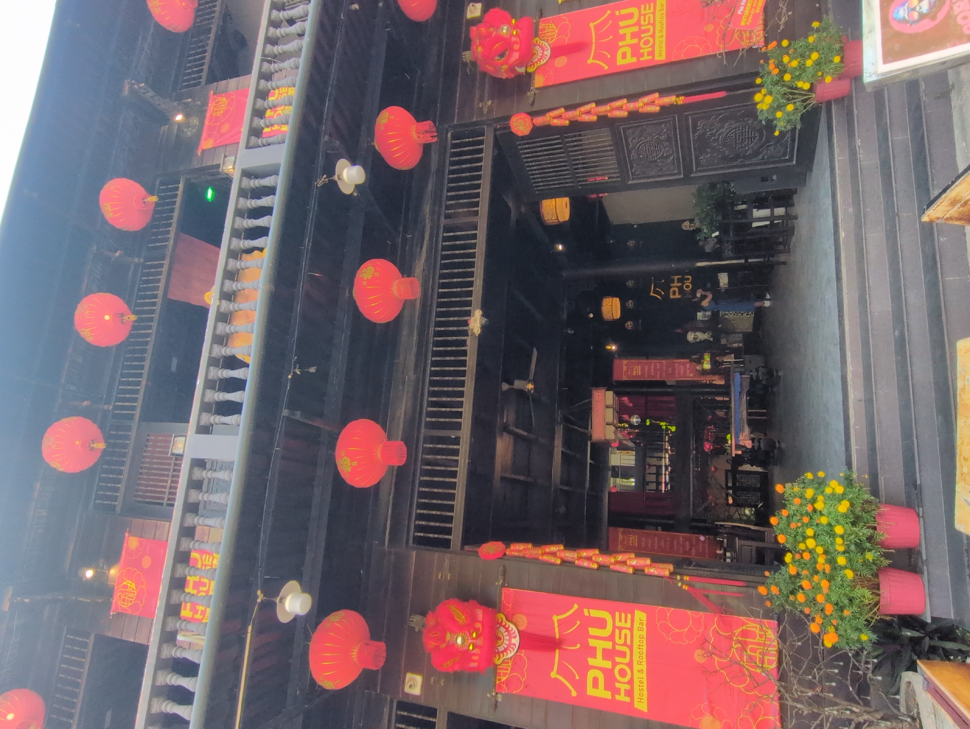 The image depicts the exterior of a hostel and rooftop bar in Phú Quốc, Vietnam. The building is decorated with red lanterns and signs with the name "PHÚ HOUSE" in yellow lettering. The building's facade is a mix of dark wood and gray concrete with architectural details such as a balcony with wooden railings. The entrance is adorned with decorative elements, including red lion figures and rows of small red lanterns strung above the door. Two potted plants with yellow and orange flowers are placed on either side of the entrance. Inside, tables and chairs can be seen, along with interior lighting. The image captures the ambiance of a commercial district at twilight.
