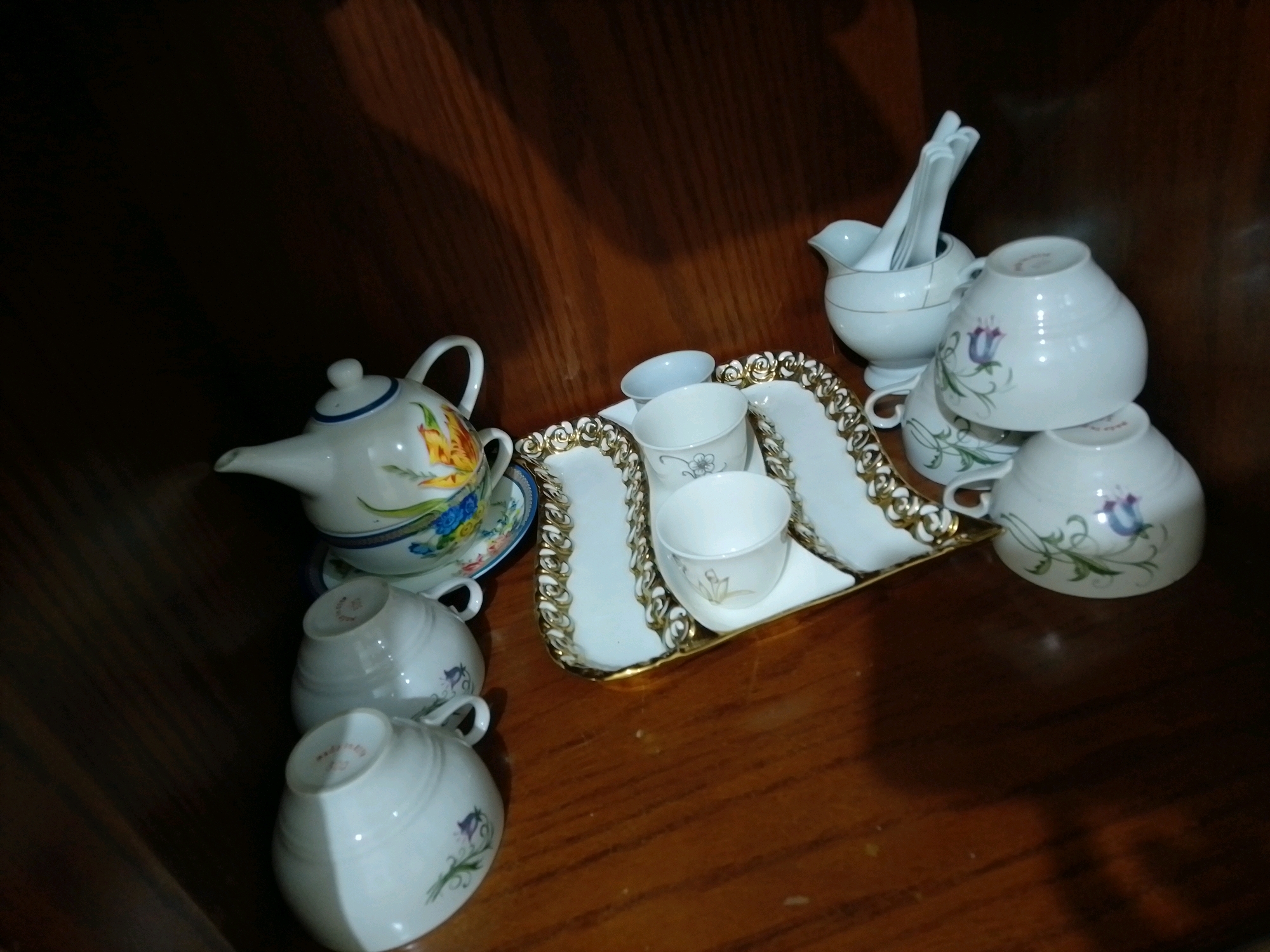 The image shows a collection of porcelain teacups and a teapot arranged on a wooden surface, possibly a table or shelf. On the left, a white teapot with a blue rim and colorful floral designs is placed on a matching saucer. To the right of the teapot, a decorative tray with a gold trim holds several white teacups, some stacked and some upright. There are also a few other white porcelain items, including what appears to be a sugar bowl with utensils. The overall scene suggests a display of fine china or a tea set. The lighting is dim, with shadows cast on the wooden surface, hinting at an indoor environment. The image does not contain any identifiable landmarks or clues to suggest a specific location within Amman, Jordan.