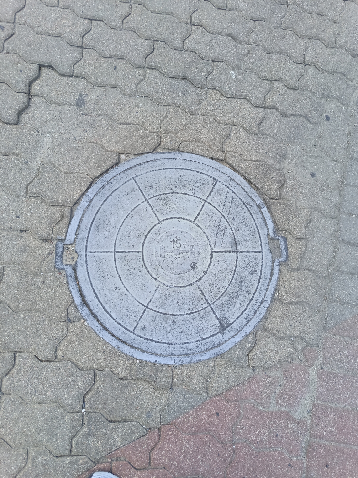 The image shows a circular manhole cover embedded in interlocking concrete pavers. The manhole cover is gray and has concentric circles and radial lines etched into its surface. In the center of the manhole cover, the number "15T" is visible, indicating its weight capacity. The pavers are a mix of gray and reddish-brown hues and show signs of wear and tear, with some cracks and missing pieces. The scene appears to be a sidewalk or pedestrian area in the city of Ivatsevichy, Belarus, as indicated by the provided location information. No people are visible in the image, and the environment is urban.
