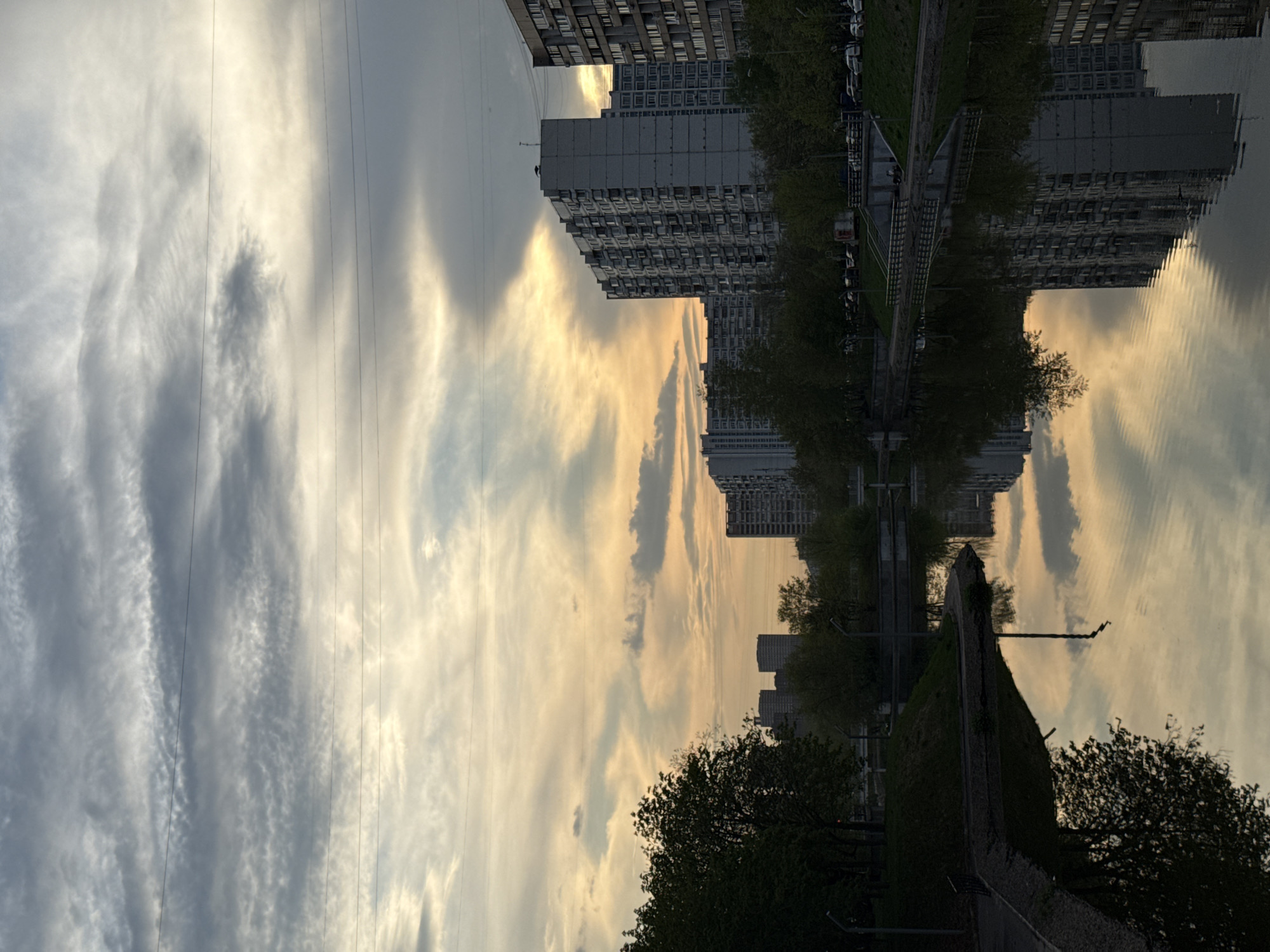A slightly rotated, wide-angle view of a cityscape with reflections on water, taken outdoors. The upper right portion shows high-rise apartment buildings with multiple windows, suggesting a densely populated urban area. Trees with sparse green foliage line the road beside the buildings, indicative of spring or early summer. The sky, taking up the upper left and central portion, shows a mix of gray and white clouds, with a touch of golden light hinting at either sunrise or sunset. Power lines stretch across the sky.
The reflection of these buildings and sky is prominently seen in a body of water, likely a river or large canal.
In the lower right, there's a view of a grassy slope with trees and a sidewalk, possibly a park area beside the water. The setting appears to be in the city.
