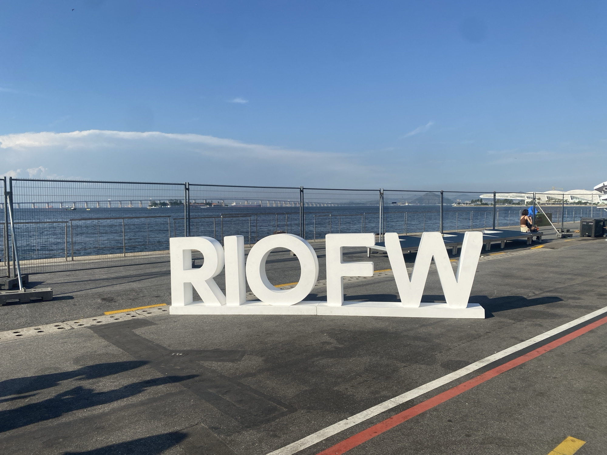 The image shows the letters "RIOFW" in large, white block letters, standing on an asphalt surface marked with red and white paint stripes. Behind the letters is a metal fence, separating the foreground from a body of water with a bridge visible in the distance. The sky is blue with some clouds. To the right of the frame, a person is sitting on a raised platform near some poles or equipment. The scene appears to be an outdoor event space located in Rio de Janeiro, Brazil.