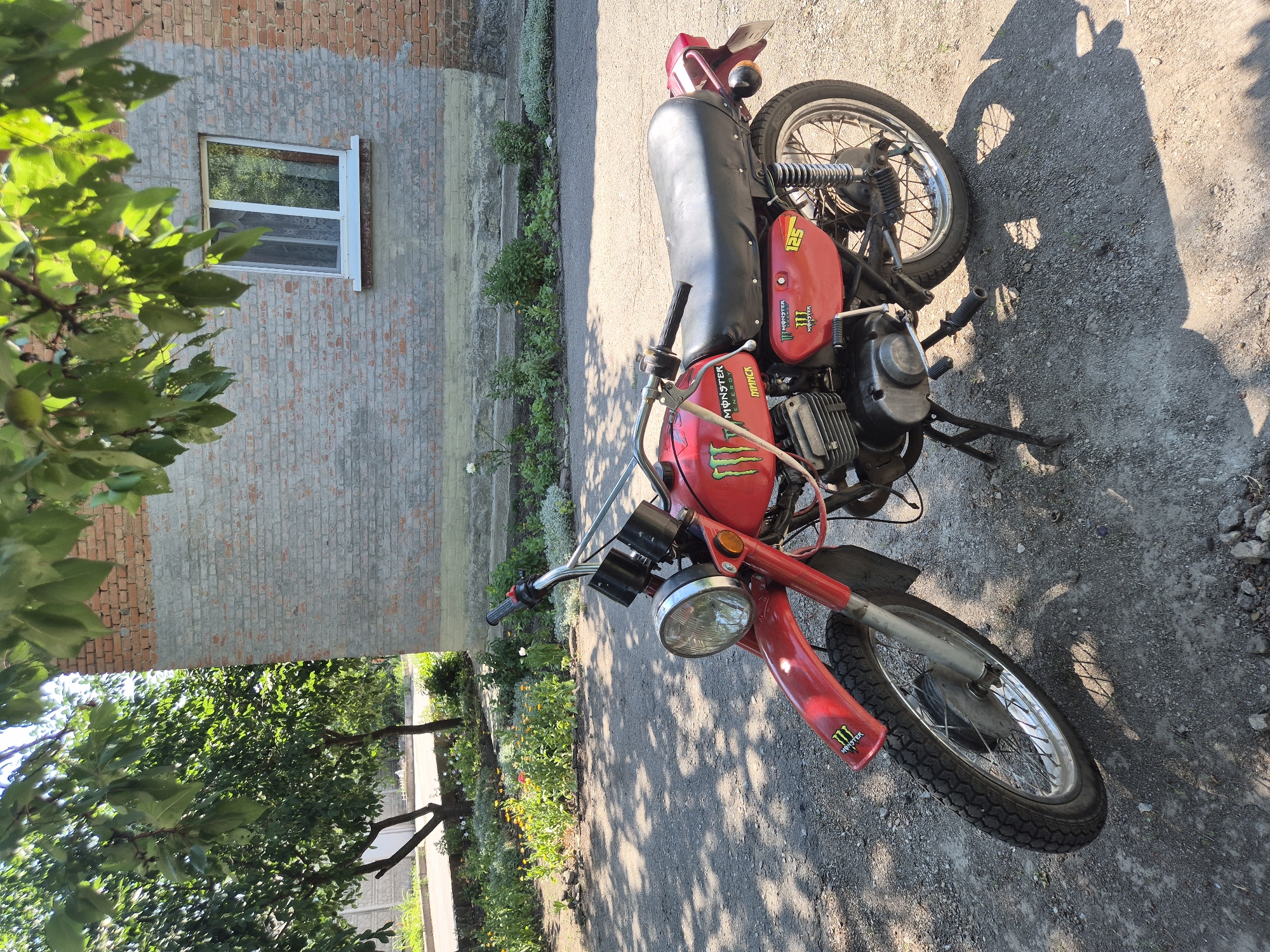 In the village of Sozonivka, Ukraine, a red vintage motorcycle with "Monster Energy" branding is parked on a gravel and asphalt surface in front of a brick building. The motorcycle is positioned at an angle, with its front wheel turned slightly to the left. It has a single round headlight, chrome handlebars, and a black seat. A kickstand supports the motorcycle. The building behind it has a light gray lower section and exposed brick above, with a white-framed window visible. Lush green trees with dappled sunlight casting shadows on the ground are present in the foreground and overhead. The surrounding area shows a flowerbed with various colorful flowers and greenery, suggesting a residential or rural setting. The sky is bright and clear, indicating a sunny day. There are no people or animals visible in the image.