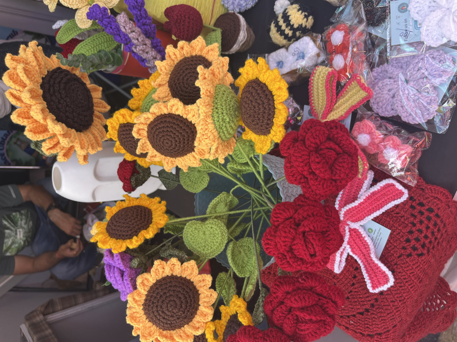 A crocheted flower arrangement, featuring sunflowers and roses, is displayed on a table in a shop in Sector 17, Chandigarh. The sunflowers are made of yellow yarn, with brown centers, and the roses are made of red yarn. The arrangement is placed in a blue bucket with green crocheted leaves, and a white plastic container sits behind it. The shop is selling other crocheted items, including a small bee, lavender flowers, and a heart-shaped crocheted blanket with a white ribbon. The scene is a close-up view of the crocheted arrangement and surrounding items. The background is out of focus.