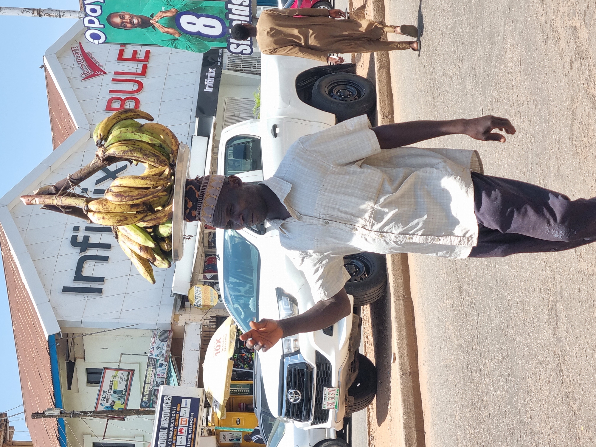 The image shows an African man balancing a tray of plantains on his head, seemingly offering them for sale. He's wearing a light-colored, button-down shirt, dark pants, and a patterned hat. He stands on a street in Jos, Nigeria, as indicated by the car's license plate "Lagos," which also displays the Nigerian flag colors. In the background are several businesses, identifiable by their signage: "Infinix," "Opay," "Buletz," and "Global Resources." A white pickup truck is parked nearby. Another man in a brown tunic stands behind the truck. The setting is an urban environment, likely a commercial district.