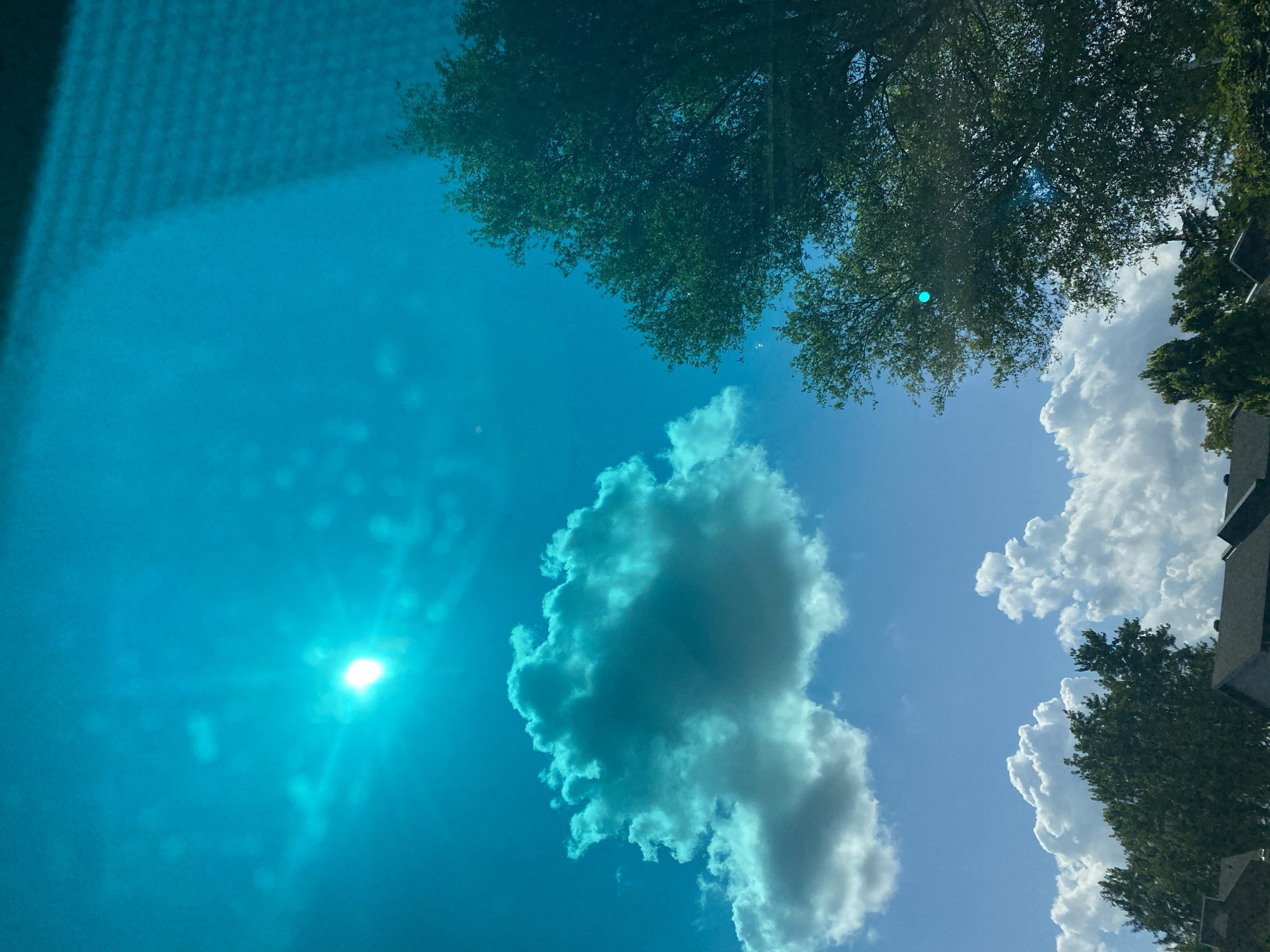 The image shows a bright sky with some clouds, trees, and rooftops in the foreground. The sky is a vibrant blue, with the sun appearing as a bright, slightly overexposed spot in the upper left quadrant. One large cloud in the center of the image has a turquoise hue, likely due to light filtering through or reflection. Several other fluffy white clouds are visible further down. Two large trees, with dense green foliage, are prominent on the right side of the image. In the lower portion, parts of several dark-colored rooftops are visible, suggesting a residential or urban area. The lighting suggests it is daytime, with the sun high in the sky. No people or animals are visible in the image. The scene appears to be a view from inside a building or vehicle, with some textural elements suggesting a window or screen in the upper right portion. The overall impression is one of a sunny day with scattered clouds and ample greenery, characteristic of a suburban or urban environment. Without specific landmarks, pinpointing the exact location within Pointe-Claire, Canada is not possible from the image alone, but it depicts a typical scene found in such a locale during daylight hours.
