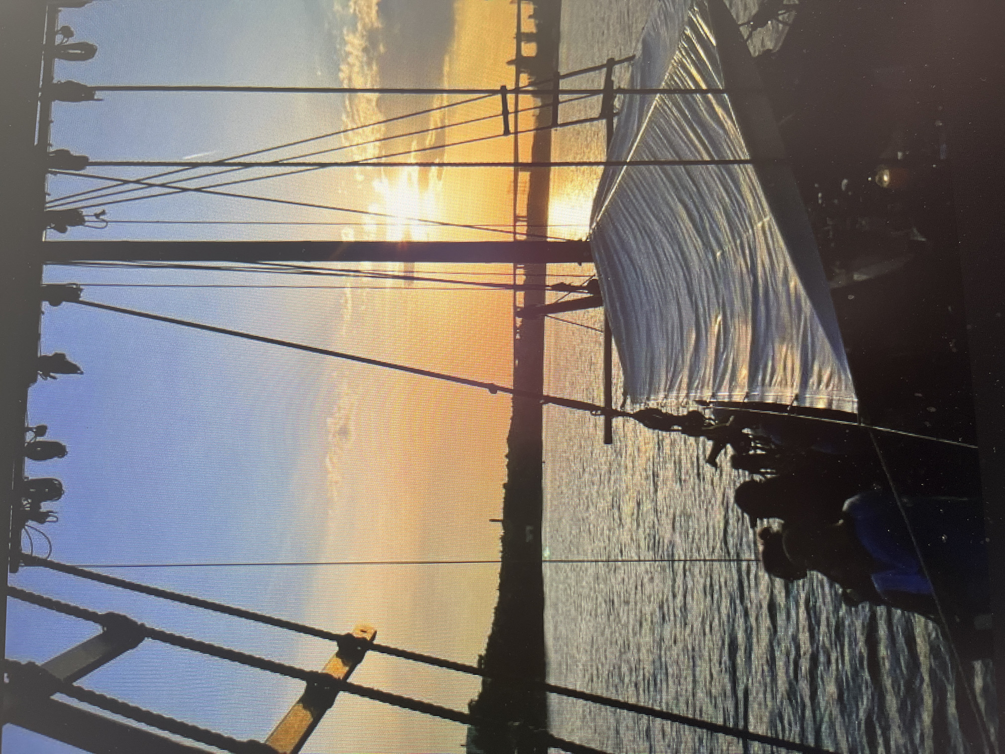 A view from a sailing ship with its sails up is shown on the water. A group of people are visible on the deck of the ship. The sky is blue to orange and the sun is setting behind what seems to be an island or a piece of land in the distance. The scene is likely taking place in the Black Sea, and its location is potentially close to Varna, Bulgaria. There is a forest and city visible on the land. There are many wires and ropes on the boat.