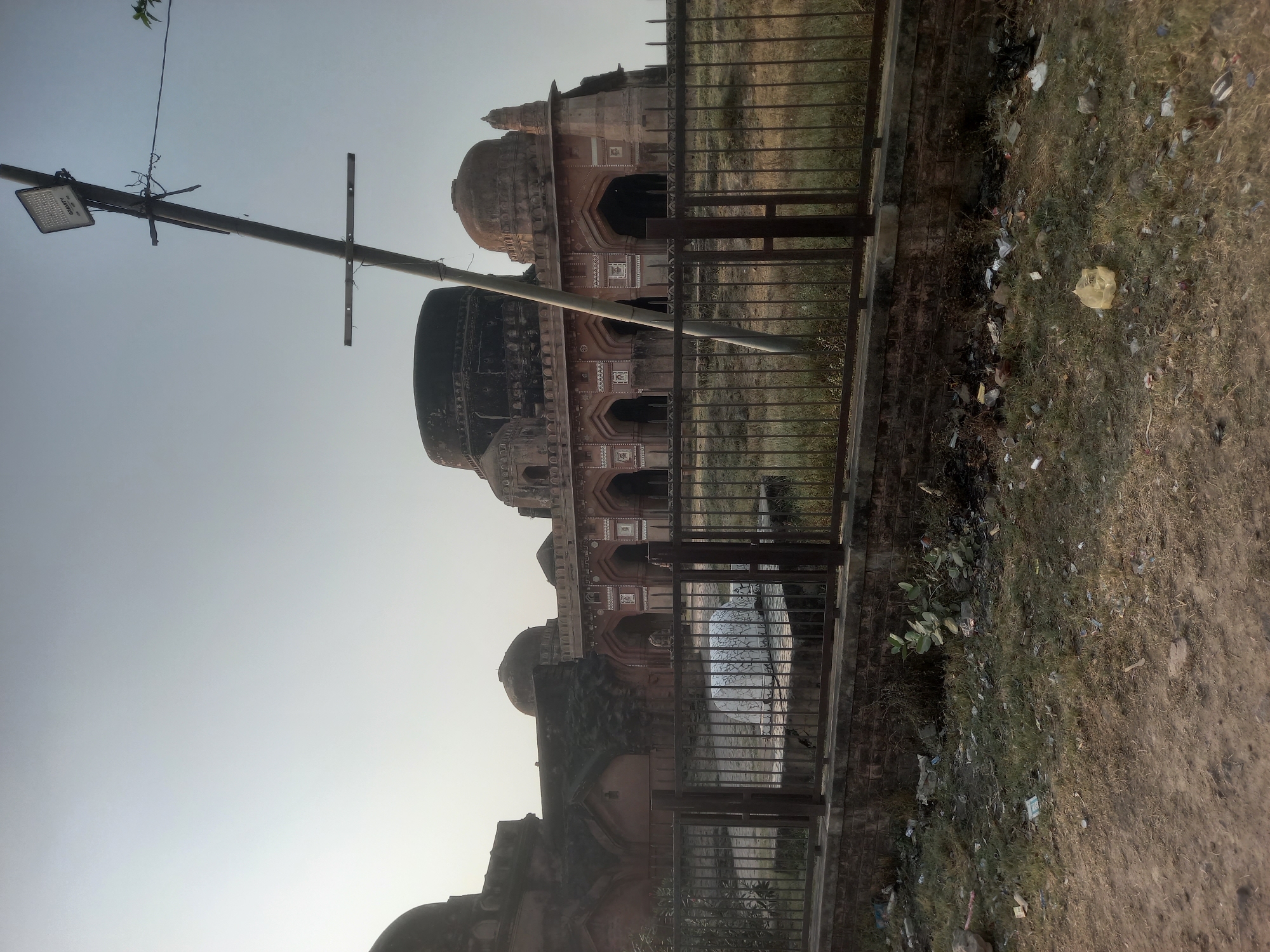 The image shows a historical building with multiple domes in Usargaon, India. A metal fence separates the building from the viewer. A light pole with a sign is visible in the foreground. The ground is covered in dirt and trash. The sky is clear and bright.