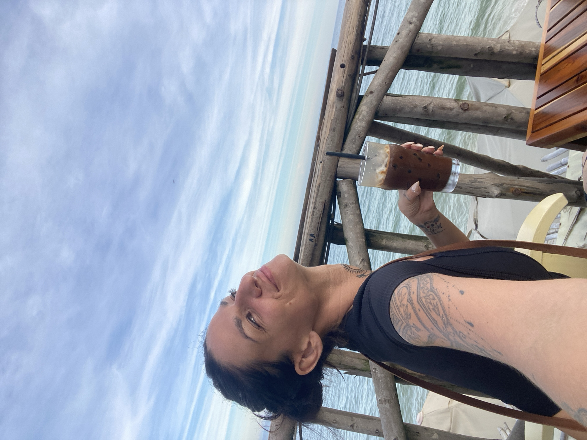 A woman with dark hair tied back, wearing a black top, is holding a clear glass with a dark beverage and a straw. She is looking up and to the side with a smile. She has tattoos on her left arm and neck. The setting appears to be an outdoor cafe with a wooden railing and furniture, overlooking the ocean. The sky is blue with wispy white clouds. The image is oriented vertically, with the sky occupying the majority of the frame. The location context is Phu Thuy Ward, Vietnam.