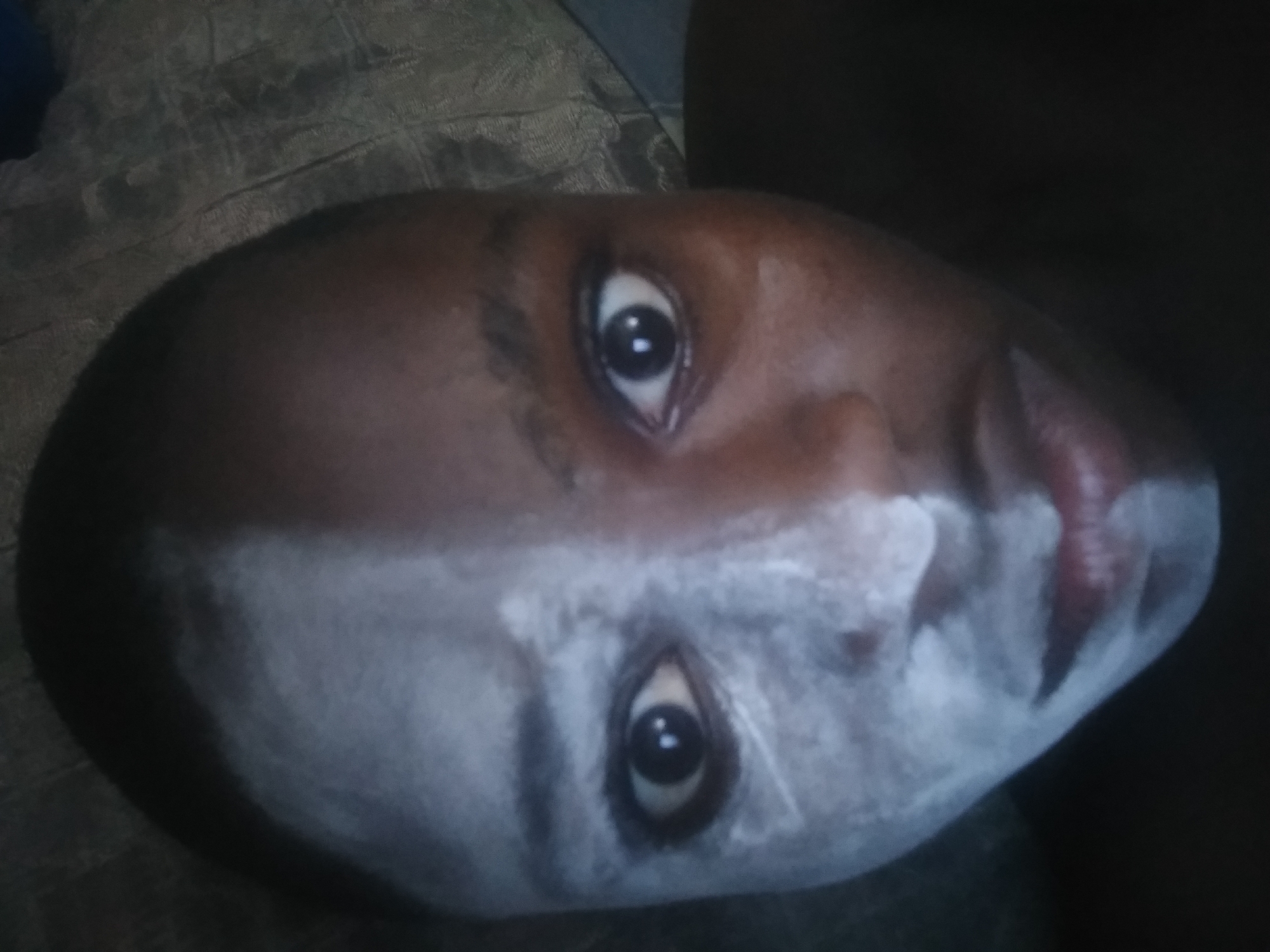 The image displays a close-up of a person's face, rotated 90 degrees clockwise. The subject appears to be a young individual with a dark skin tone. Their face is divided into two distinct sections by a thick, uneven layer of white substance. The upper part of the face, including the forehead, eyebrows, and the area around the top of the eyes, is visible in its natural dark complexion. The lower part of the face, encompassing the cheeks, nose, mouth, and chin, is heavily covered in the white material.
Both eyes are wide open, with prominent white sclera and large, dark pupils, fixed in an intense, direct gaze towards the viewer. The white substance creates a sharp horizontal line across the face, running just below the eyes and across the bridge of the nose. The lips, appearing reddish, are faintly visible through the white layer. The background is dark and textured, possibly a fabric surface, suggesting an indoor setting. The lighting is dim, which enhances the stark contrast between the dark skin and the bright white substance. No specific activity is depicted other than the person presenting their face with the applied white material, which could be face paint or a mask. There is no visible text in the image.