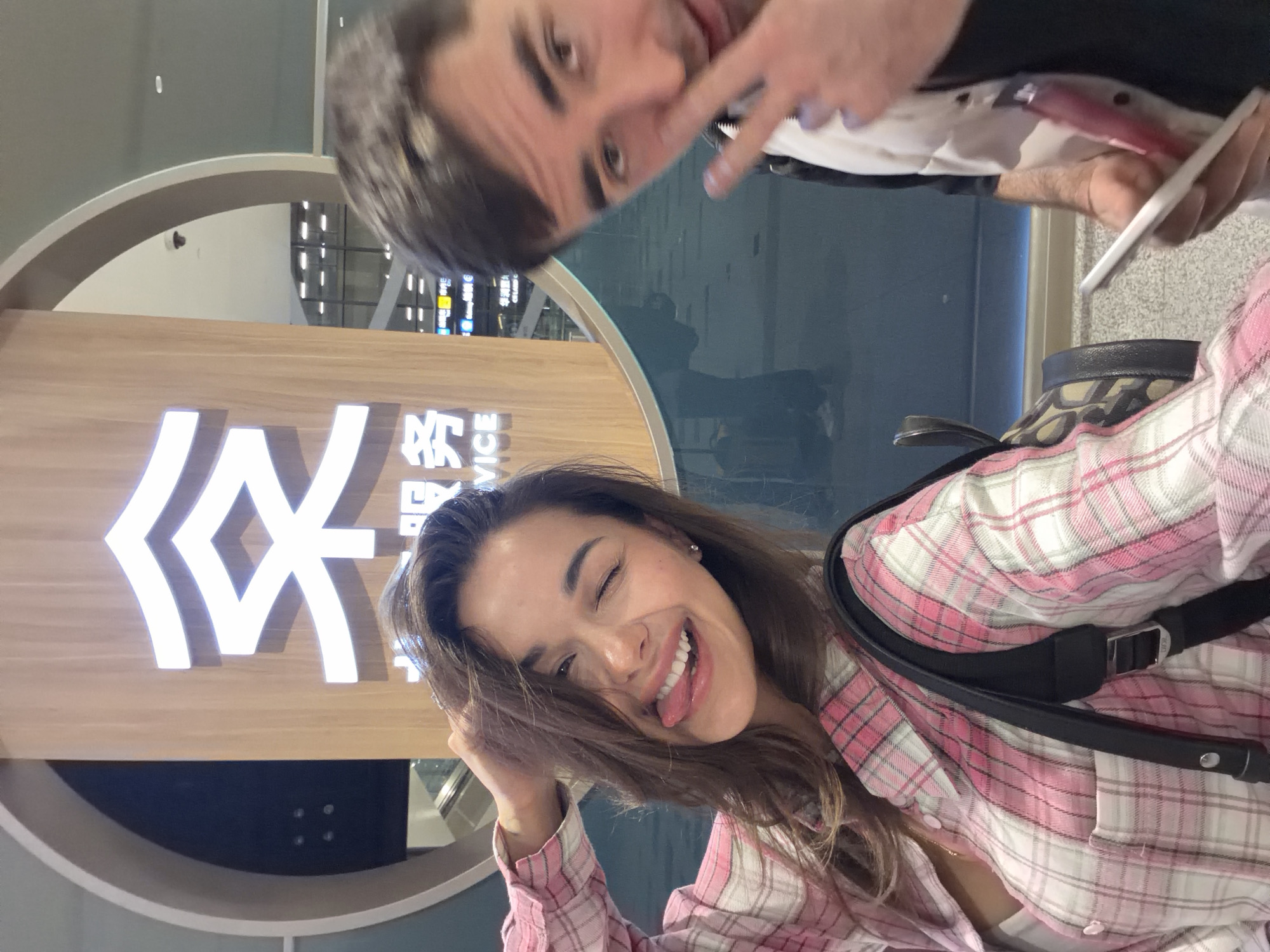 The image shows two individuals in what appears to be an airport terminal. The woman in the foreground is wearing a pink and white plaid shirt and a black backpack. She is sticking out her tongue and smiling at the camera. A man is partially visible in the background, holding up two fingers in a peace sign near his eye. They are posing in front of a sign that reads "SERVICE" with Chinese characters above it. The backdrop suggests a modern airport setting with glass walls and informational signage. The location is identified as Langfang, China.