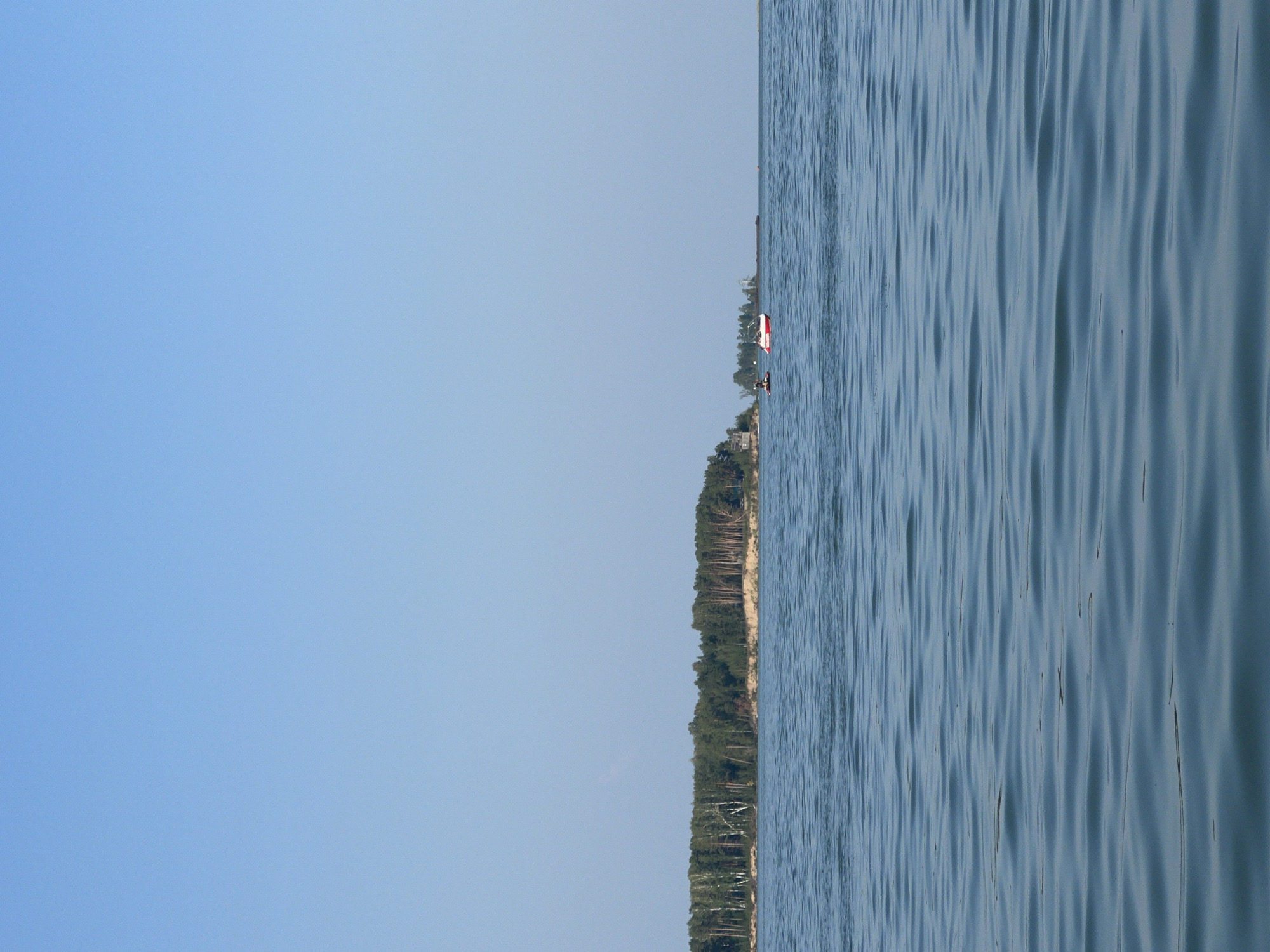 This is a wide-angle, eye-level, outdoor, daytime photograph of a wide body of water under a clear blue sky. The water is calm with slight ripples and reflects the sky. On the left side of the image, a densely wooded shoreline with sandy patches is visible. A few structures, possibly houses, are nestled amongst the trees on the shoreline. In the middle distance, a red and white boat is moving on the water, accompanied by a dark-colored object that might be another small boat or a person on a paddleboard. Further to the right, a long, dark, barge-like object is also visible on the water, and beyond that, a thin strip of land or a distant shoreline. The overall scene depicts a serene, natural landscape.