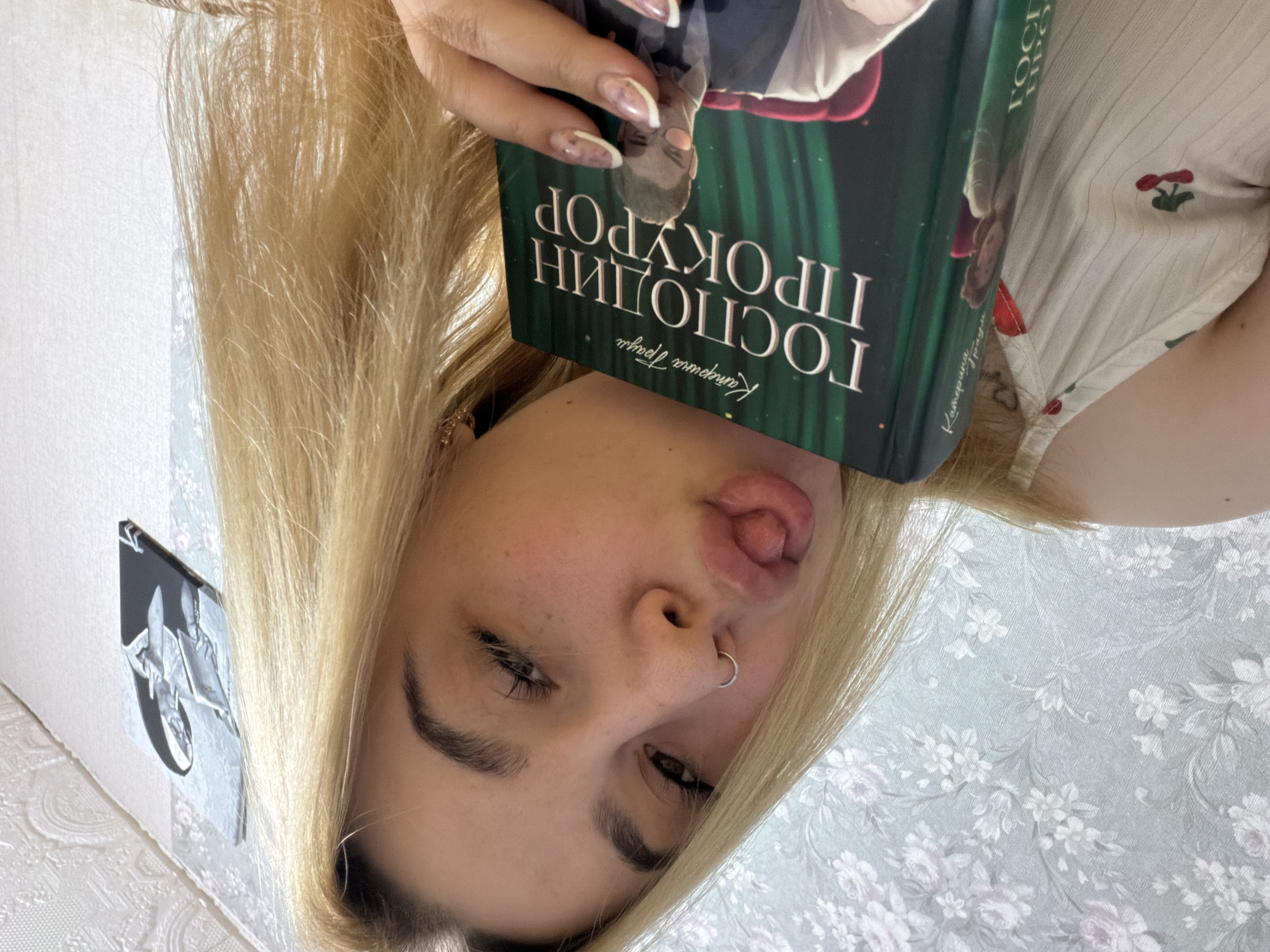 A young woman with blonde hair is holding a book in front of her face. She is sticking her tongue out playfully. The book has text in Cyrillic, and the title appears to be "ГОСПОДИН ПРОКУРОР" (Mr. Prosecutor). The woman has a nose ring in her left nostril and manicured nails. The background shows a floral patterned wallpaper and a black and white artwork on the wall. The image was likely taken indoors. There is no information to verify the specific location as Chaikovsky, Russia.