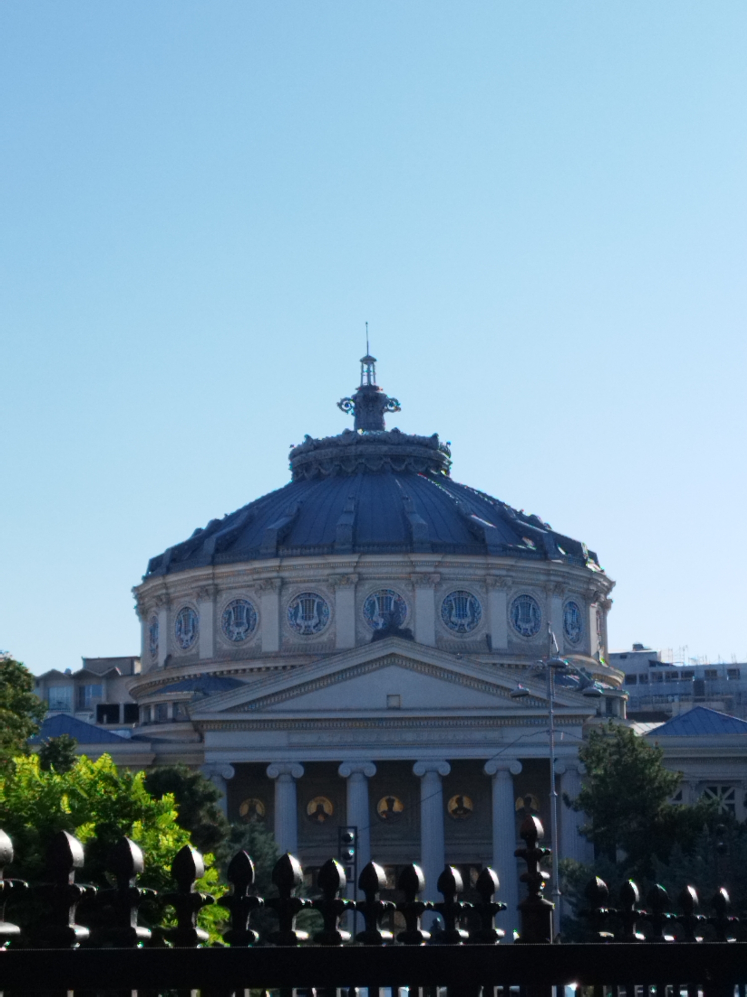 The image shows the Romanian Athenaeum in Bucharest, Romania, a landmark neoclassical concert hall. The building features a prominent dome and a portico with columns. In the foreground, a black wrought-iron fence is visible, with leafy green trees behind it. The sky is clear and blue. The image is captured on a sunny day, with the building and its surroundings bathed in natural light.