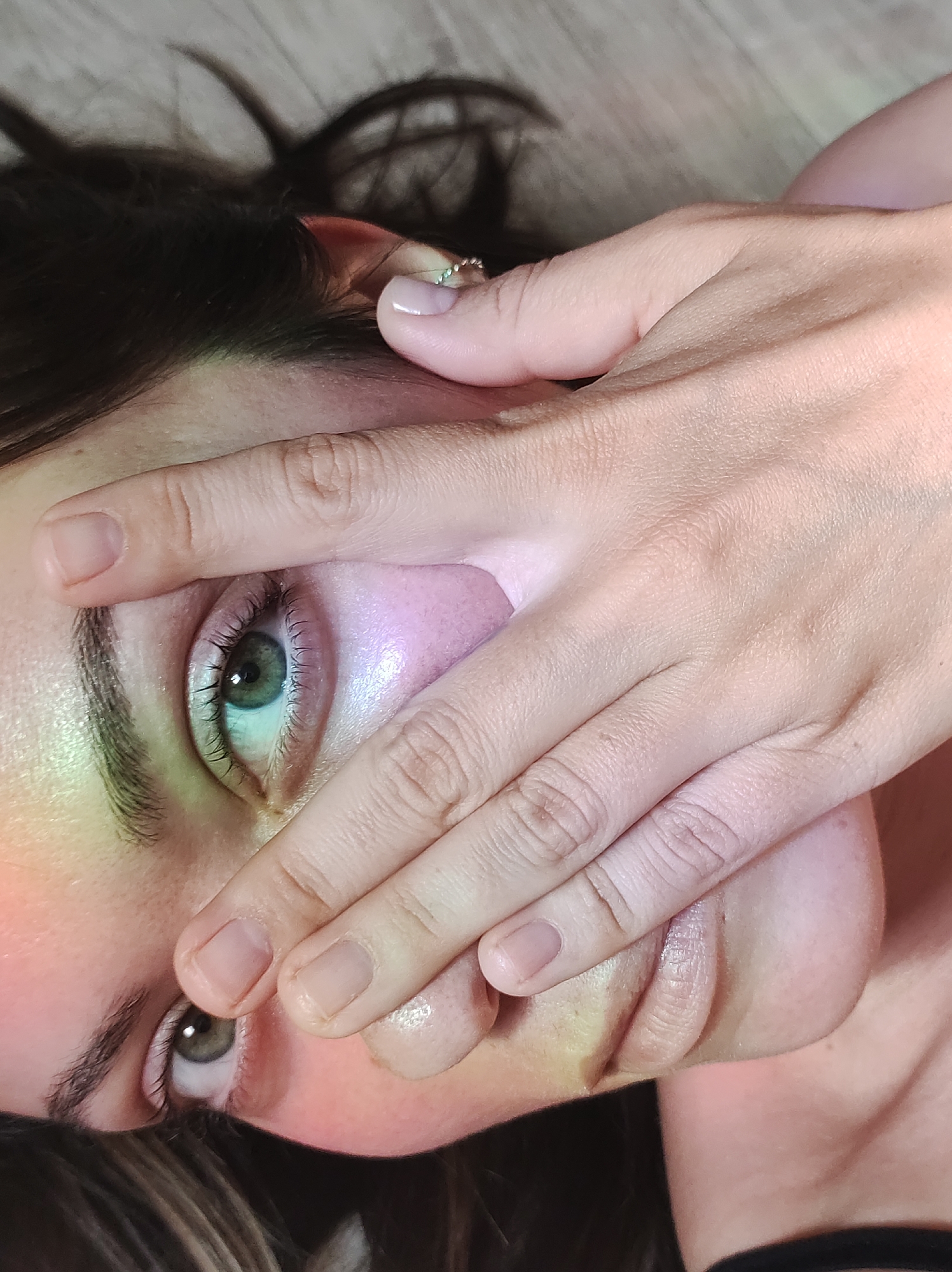 A close-up, angled view shows a person with a hand partially covering their face. The person has fair skin, brown hair, and green eyes. The hand is positioned over the face from the forehead down to the mouth. The fingernails appear to be unpolished. The image is not set in any specific location.