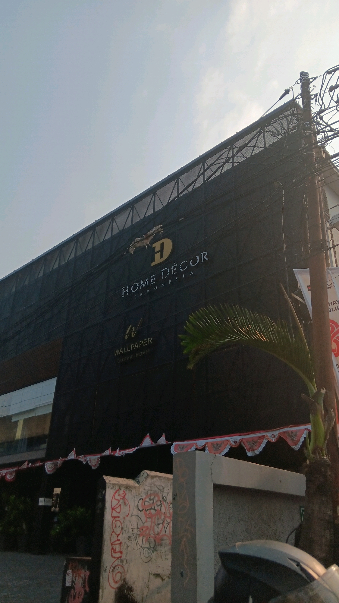 A low-angle, wide shot captures the facade of a building in South Jakarta, Indonesia, under a clear sky. The building is mostly covered by black scaffolding, with the words "HOME DÉCOR INDONESIA" and "WALLPAPER GRAHA INDAH" visible in gold and white lettering. A palm frond extends into the frame from the right side, partially obscuring the building. Below the building, a concrete wall is covered in red and black graffiti. A string of red and white pennants hangs along the base of the building, and a portion of a vehicle is visible in the foreground on the right. A utility pole with numerous wires stands to the right of the building.